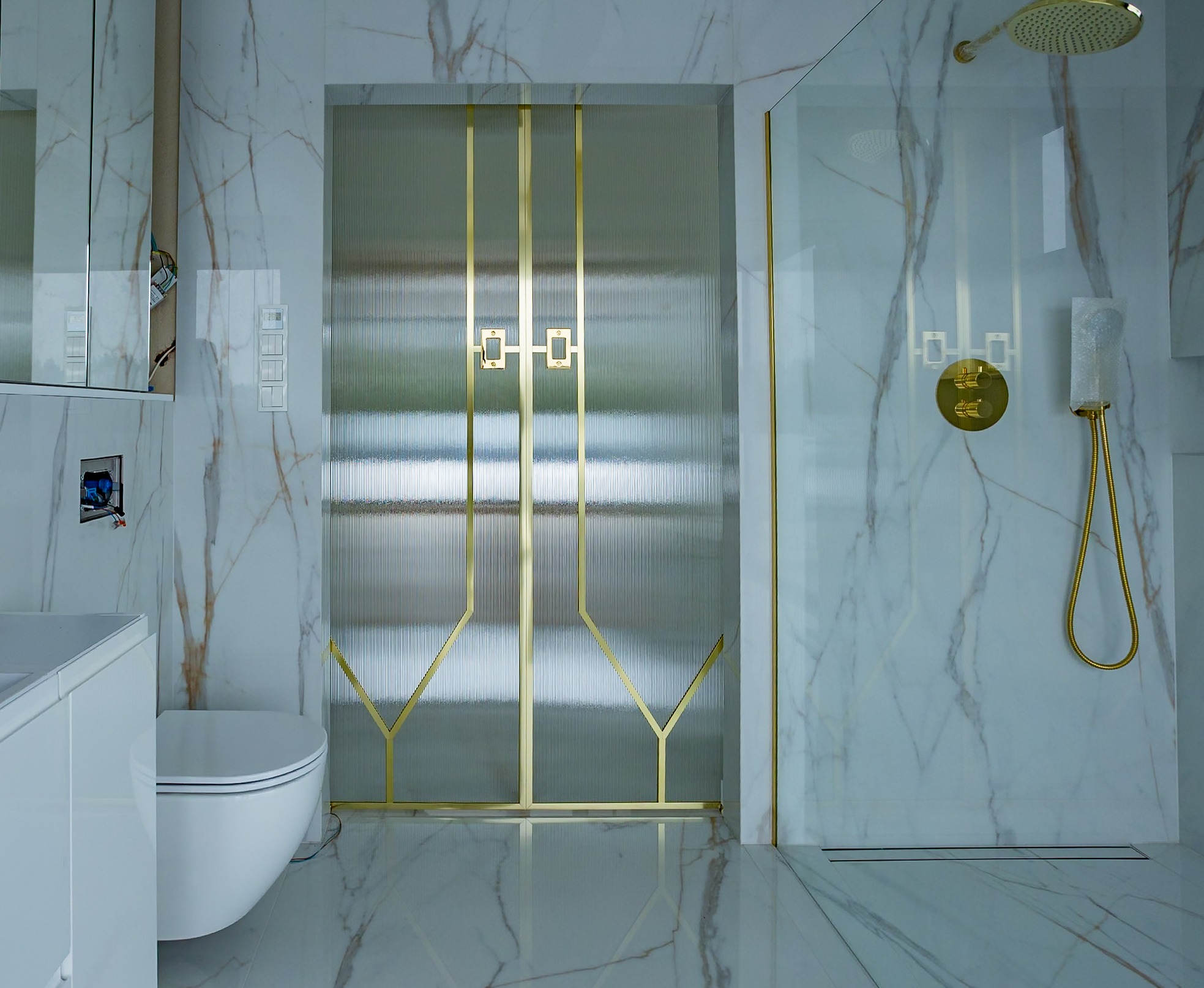 Nowoczesna łazienka z kabiną prysznicową ze szkła ryflowanego i złotymi detalami. Marmurowe płytki na ścianach i podłodze. Widoczna armatura w kolorze złota.