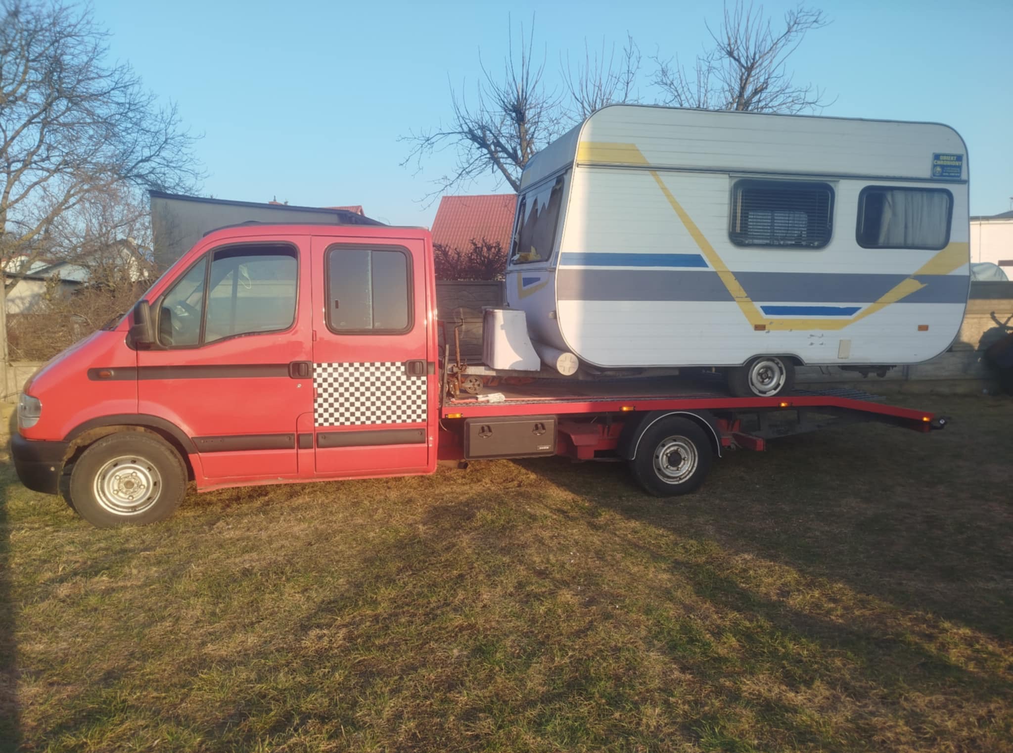 Czerwona laweta z szachownicą na drzwiach transportuje przyczepę kempingową na zewnątrz, na trawiastym terenie, w słoneczny dzień.