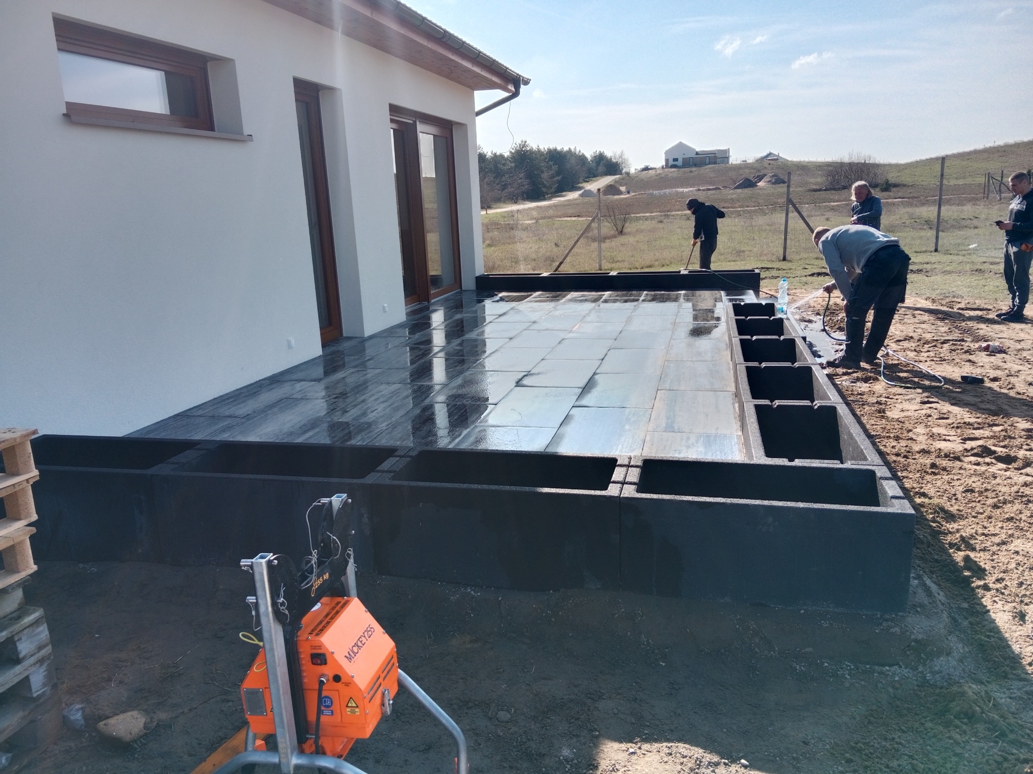 Montaż tarasu z ciemnej kostki brukowej przy domu. Widoczna ubijarka, pracownicy podczas czyszczenia powierzchni. W tle zabudowa i pagórkowaty teren.