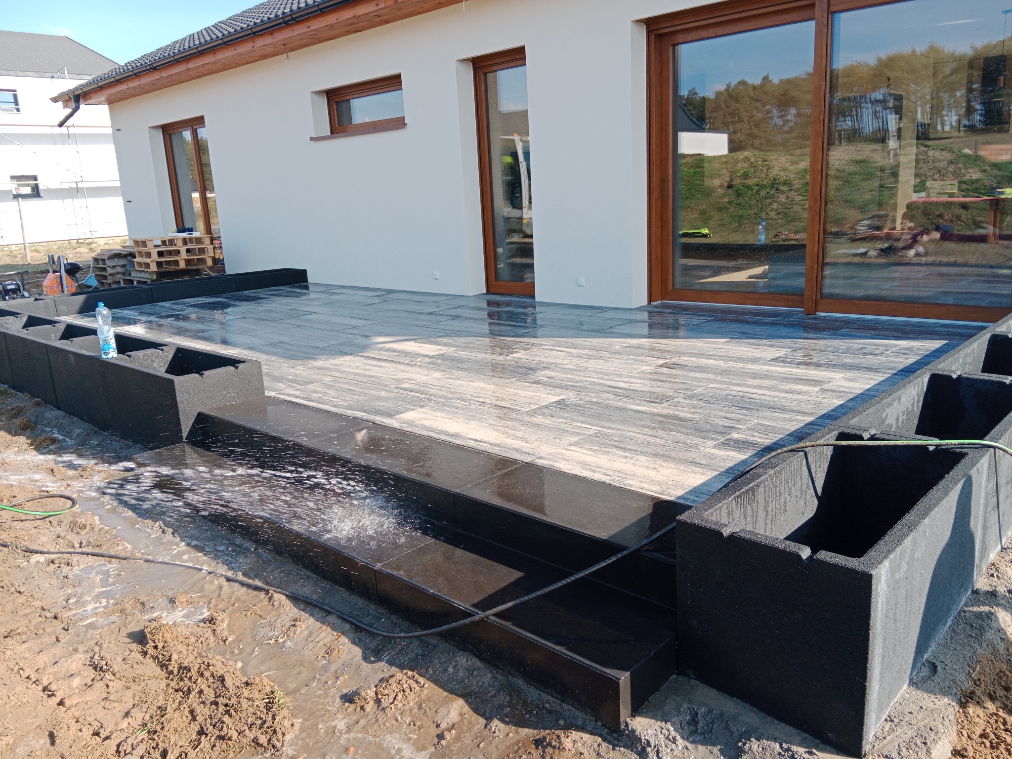 Nowoczesny taras z szarych płytek i czarnych obrzeży, myty pod ciśnieniem. Widoczne duże okna domu i przygotowane rabaty na rośliny.