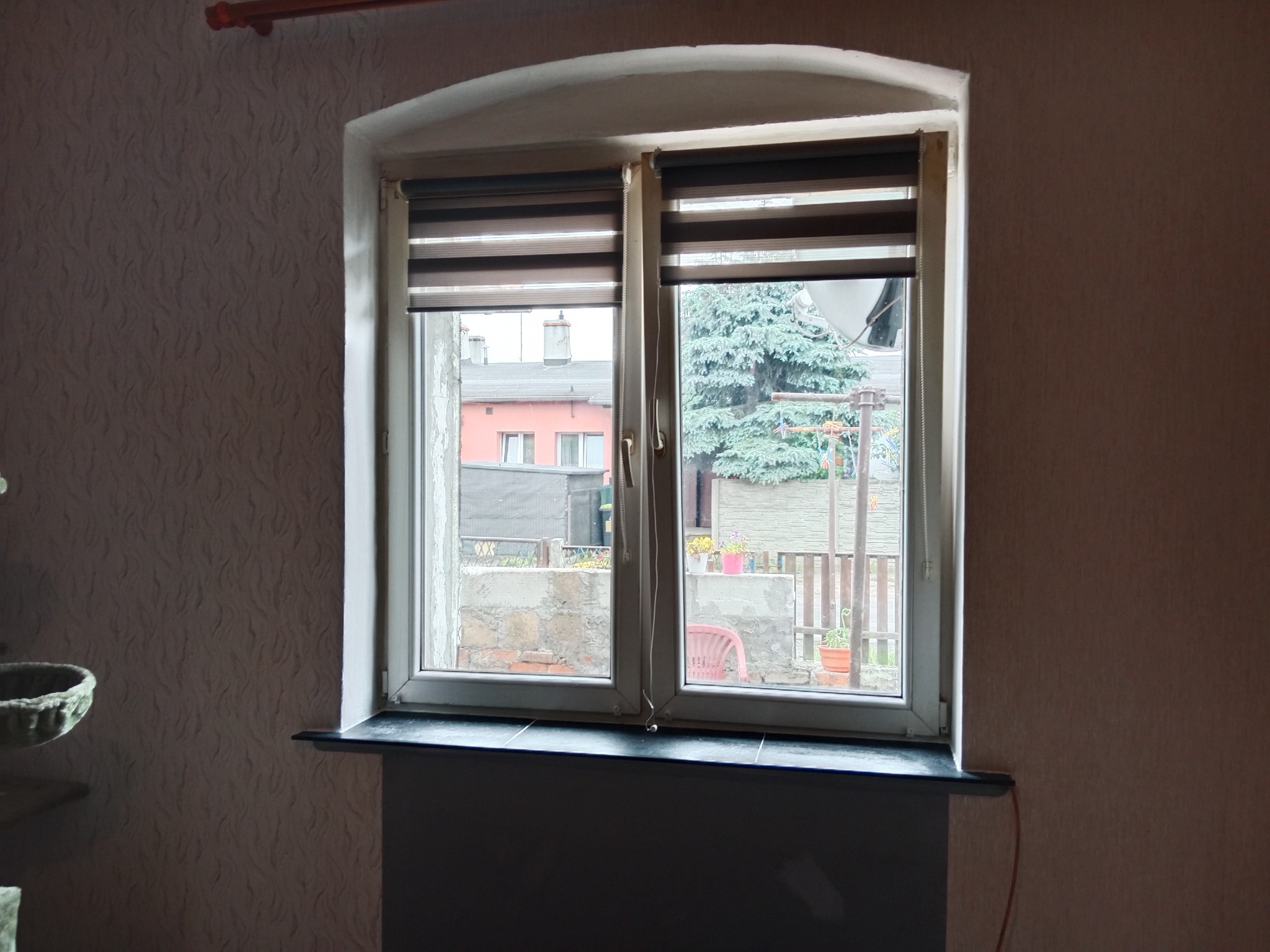 Okno z roletami dzień-noc w białej ramie, widok na podwórko z zielenią i budynkami. Dekoracyjna tapeta na ścianie.