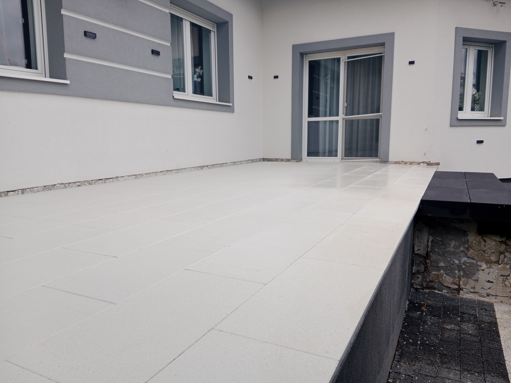 Jasne, prostokątne płyty tarasowe ułożone na zewnątrz budynku z szarymi oknami i drzwiami przesuwnymi. Widoczny fragment obniżonego, czarnego tarasu.