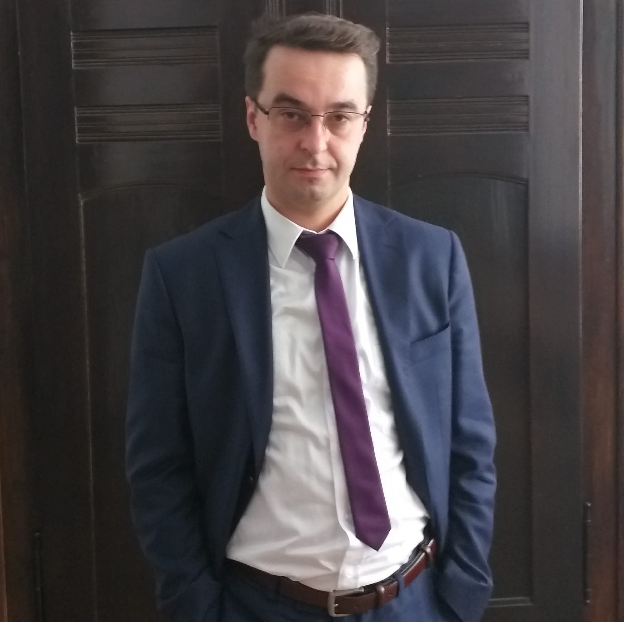 Portret adwokata w Bolesławcu w niebieskim garniturze i fioletowym krawacie, stojącego na tle ciemnych drzwi. Mężczyzna w okularach patrzy wprost w obiektyw.