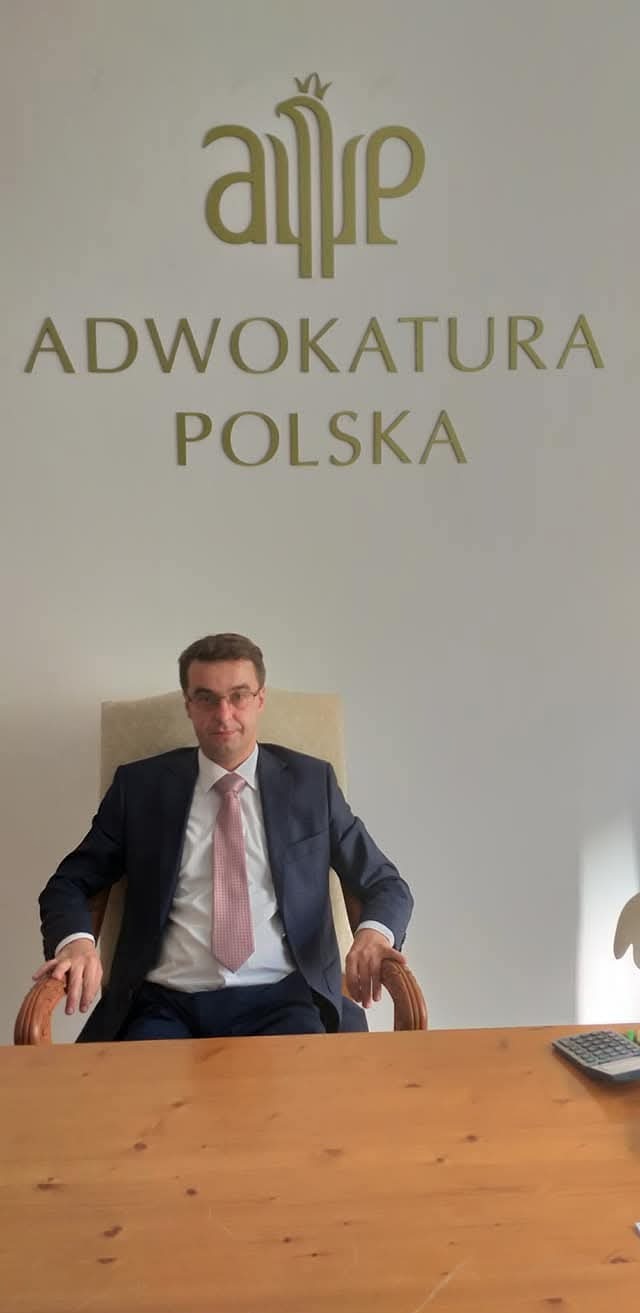 Portret adwokata w biurze pod znakiem Adwokatury Polskiej. Mężczyzna w garniturze siedzi za biurkiem, w tle logo kancelarii.
