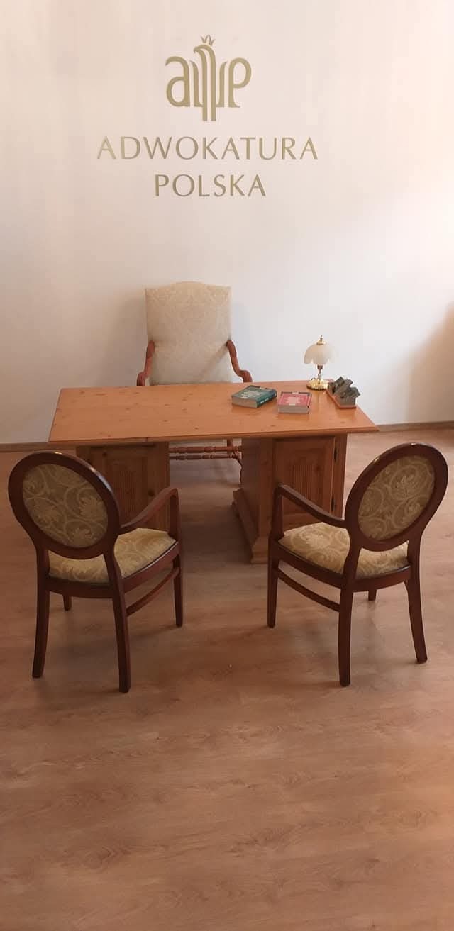 Jasne biuro adwokackie z drewnianym biurkiem, krzesłami dla klientów i logo 'Adwokatura Polska' na ścianie. Na biurku lampka i książki.
