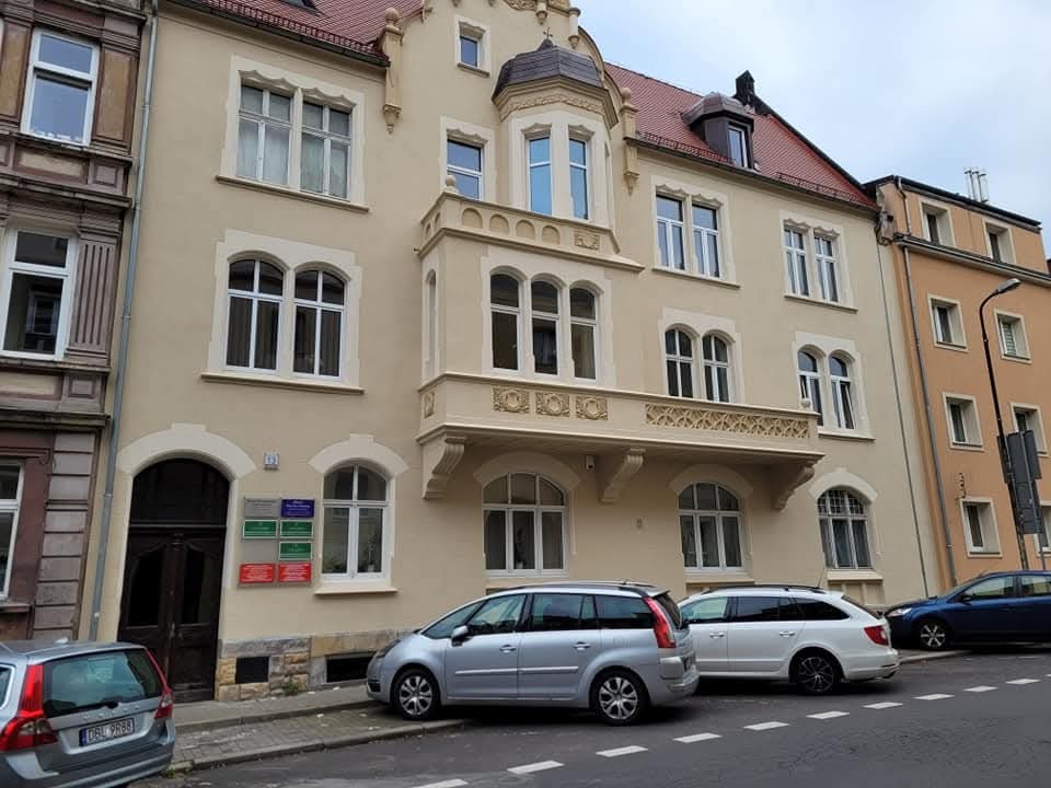 Elewacja kamienicy z balkonem i tabliczkami informacyjnymi przy wejściu. Przed budynkiem zaparkowane samochody. Widok z ulicy, pochmurne niebo.