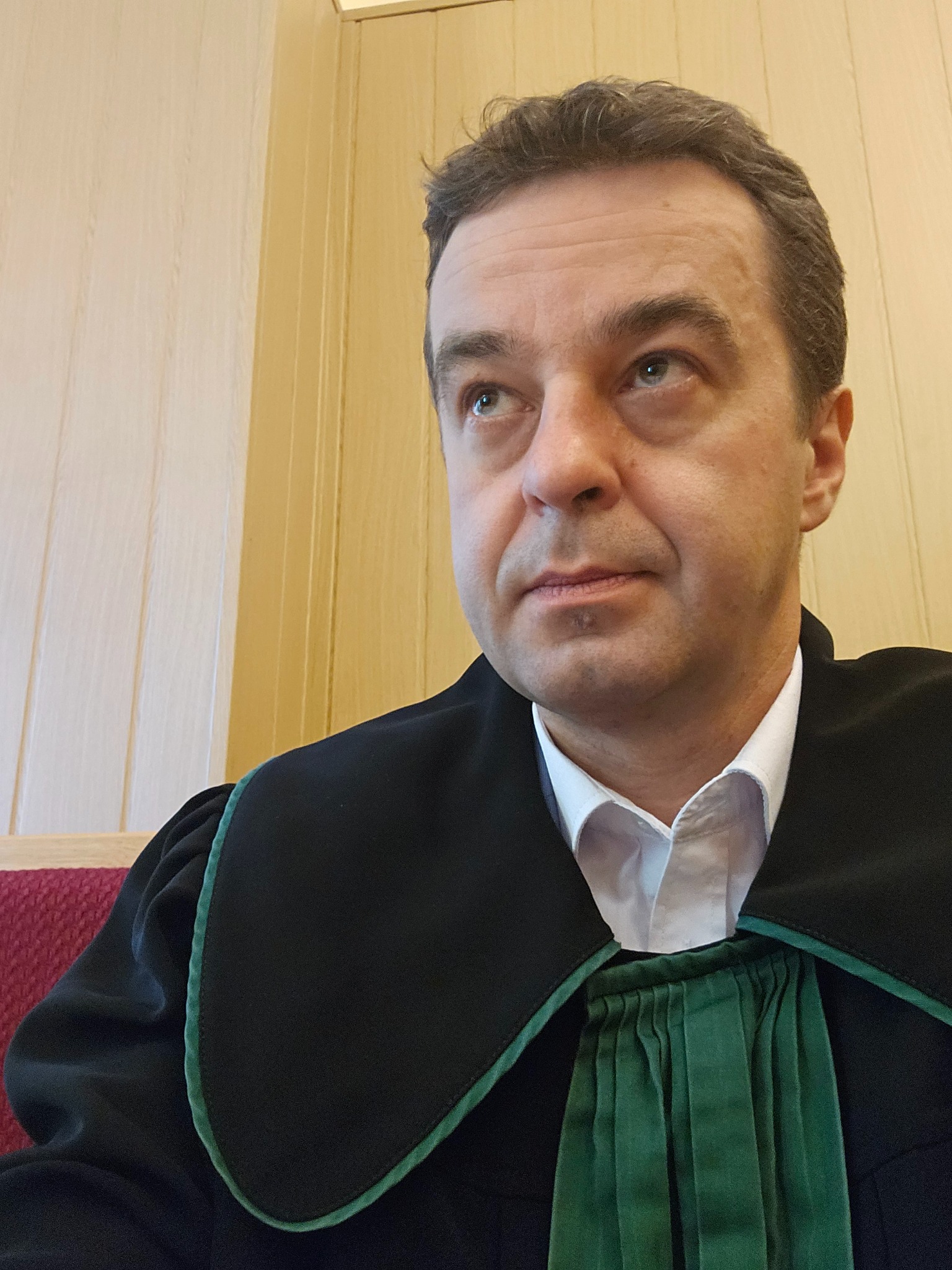 Portret mężczyzny w todze adwokackiej, spoglądającego w górę. Widoczne detale togi i fragment wnętrza sali sądowej.