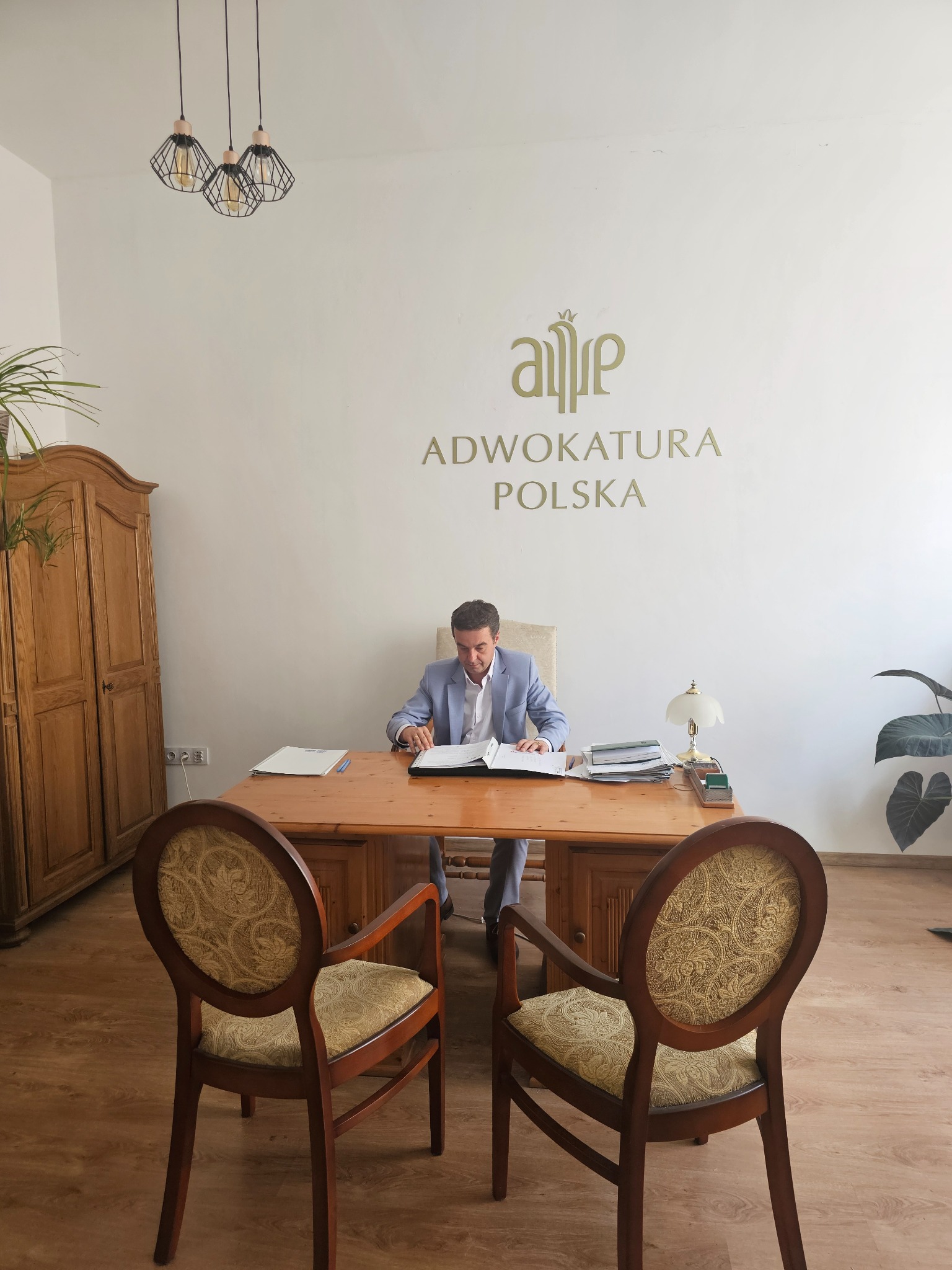 Adwokat w jasnym garniturze siedzi przy drewnianym biurku w kancelarii z logo 'Adwokatura Polska' na ścianie. Dwie krzesła dla klientów stoją przed biurkiem.