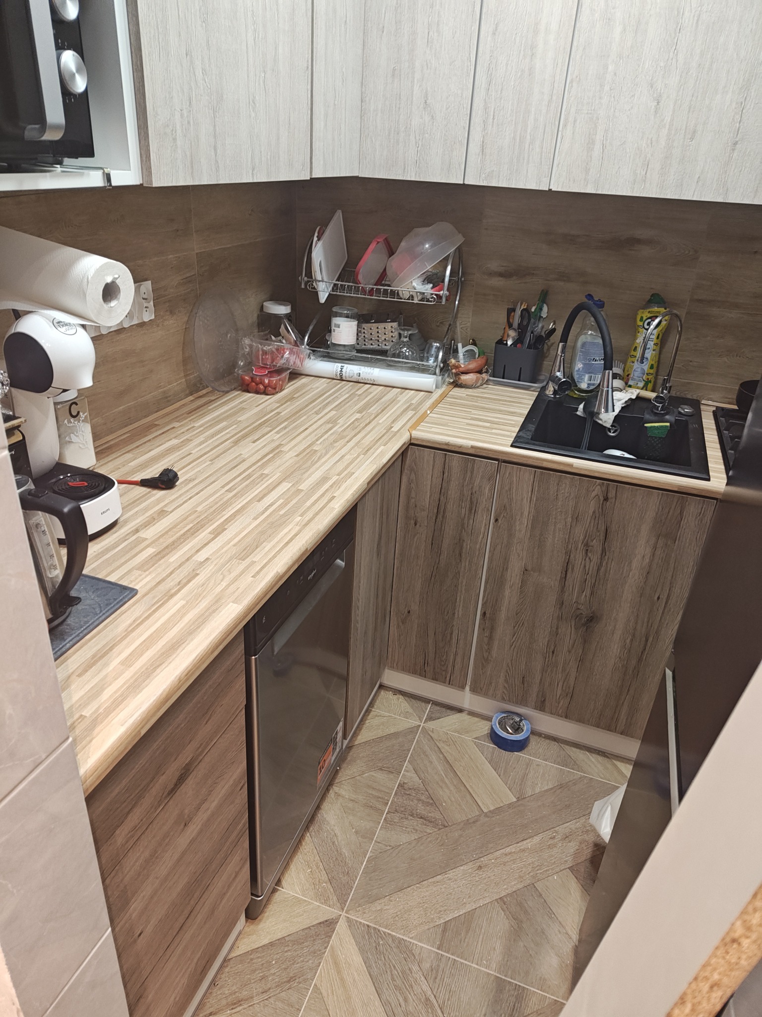 Nowoczesna kuchnia w kształcie litery L z drewnianymi frontami, blatem imitującym drewno i czarnym zlewozmywakiem. Widoczny ekspres do kawy i akcesoria kuchenne.