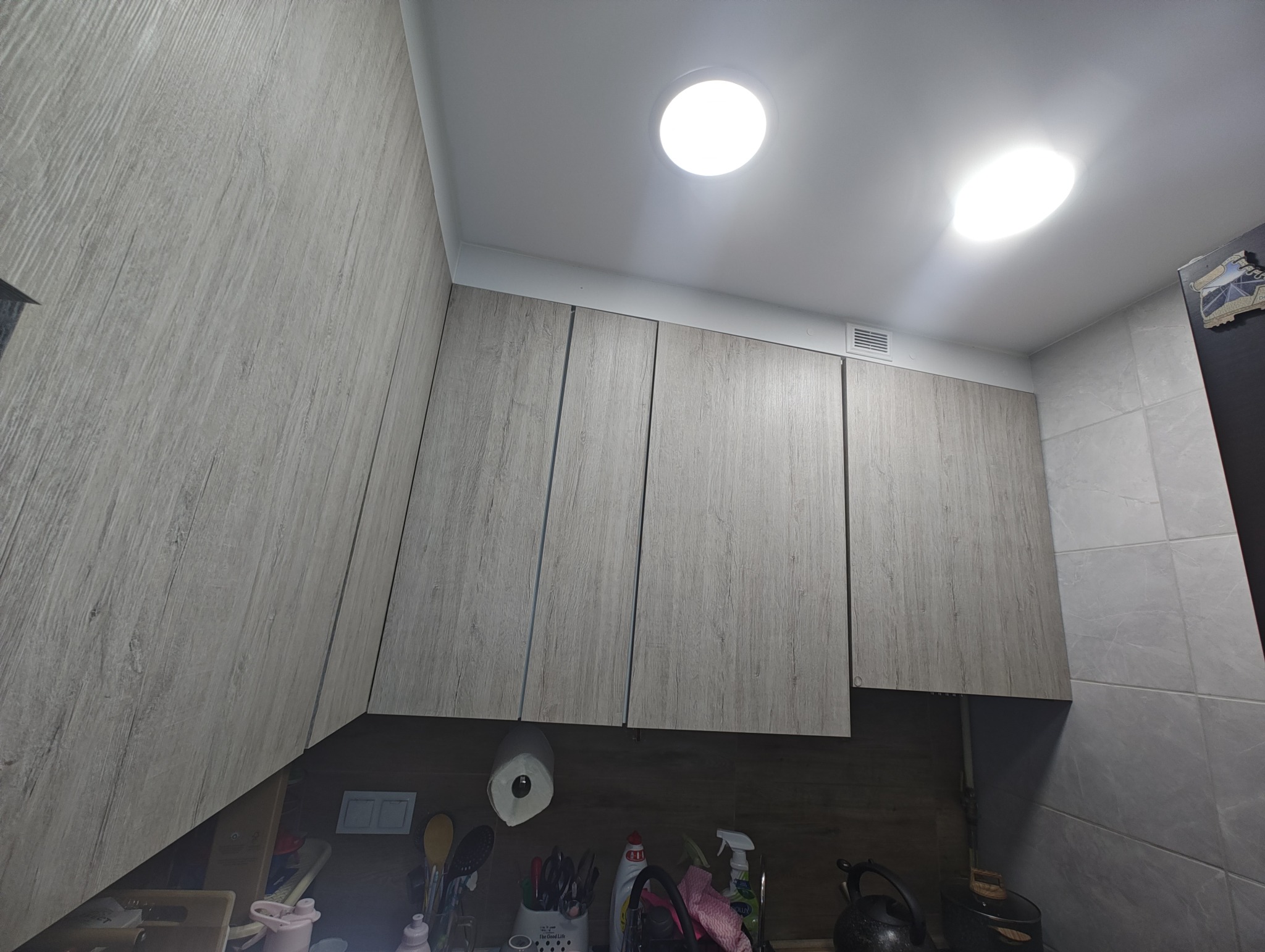 Szare szafki kuchenne z drewnianą fakturą pod sufitem, oświetlone okrągłymi lampami. Widoczny fragment ściany z szarymi płytkami. Ujęcie z dołu.