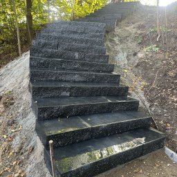 Slawomir Gardjas - Granitowe schody na zboczu, prowadzące przez las. Widoczne czarne, błyszczące stopnie i surowe, ziemne otoczenie. U góry sylwetka osoby.