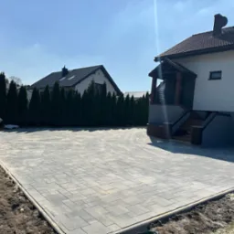 Szeroki podjazd z szarej kostki brukowej ułożonej we wzór prostokątów, obramowany obrzeżem. W tle dom z brązowym dachem i rząd tuj. Słoneczny dzień.