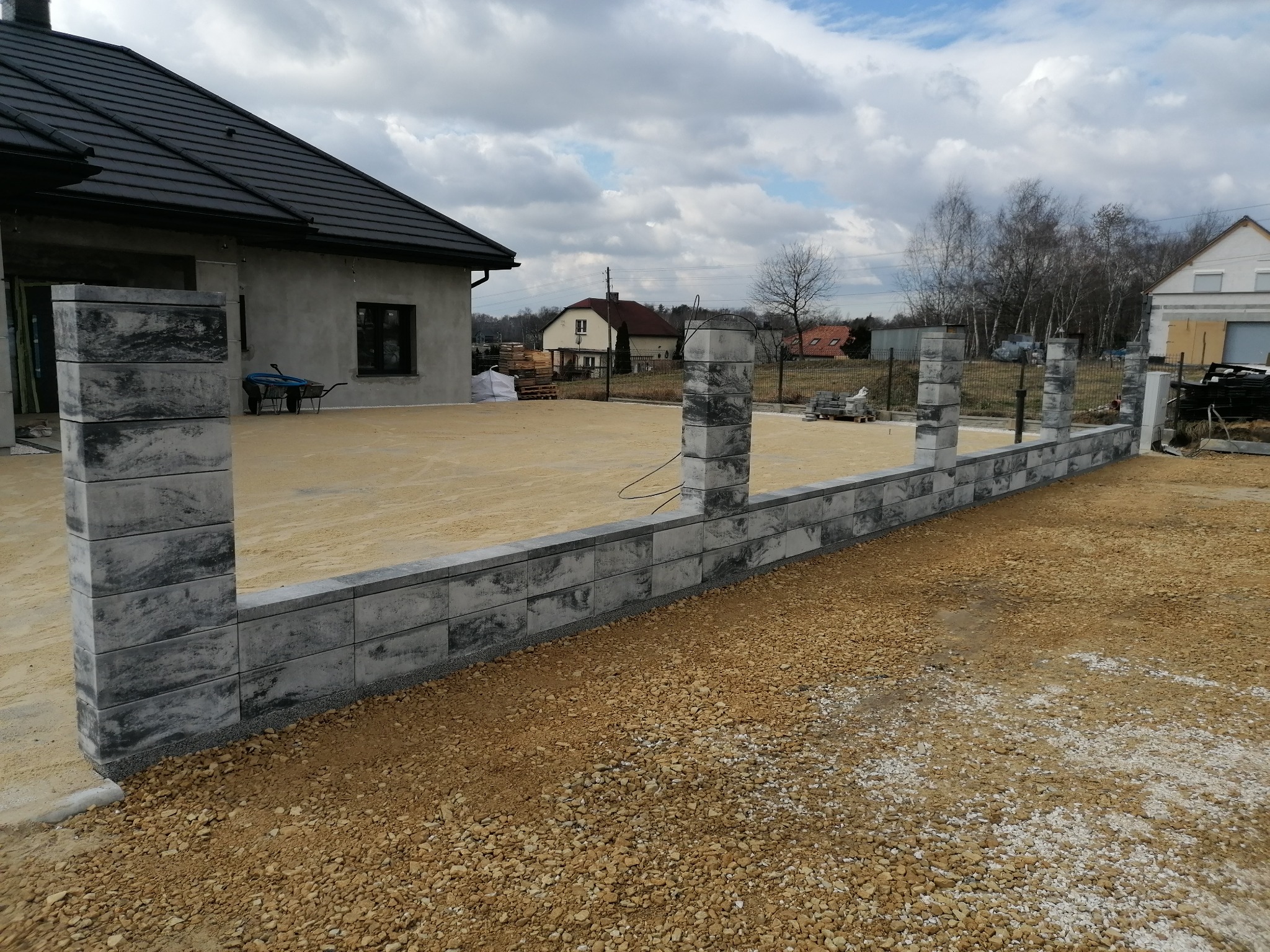 Wzniesiony fragment muru z szarych bloczków betonowych, widoczny na tle placu przygotowanego pod budowę. W tle dom jednorodzinny i pochmurne niebo.