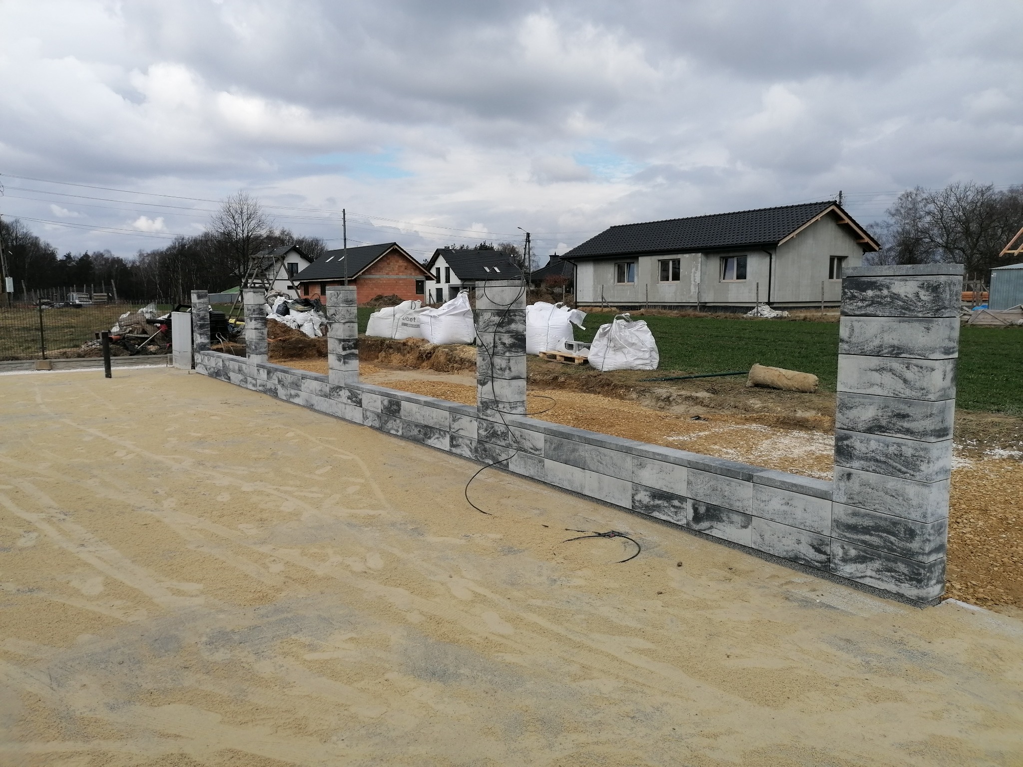 Wznoszona z szarych bloczków ogrodzeniowych podmurówka na przygotowanym podłożu. W tle budynki mieszkalne i worki z materiałem budowlanym. Pochmurne niebo.