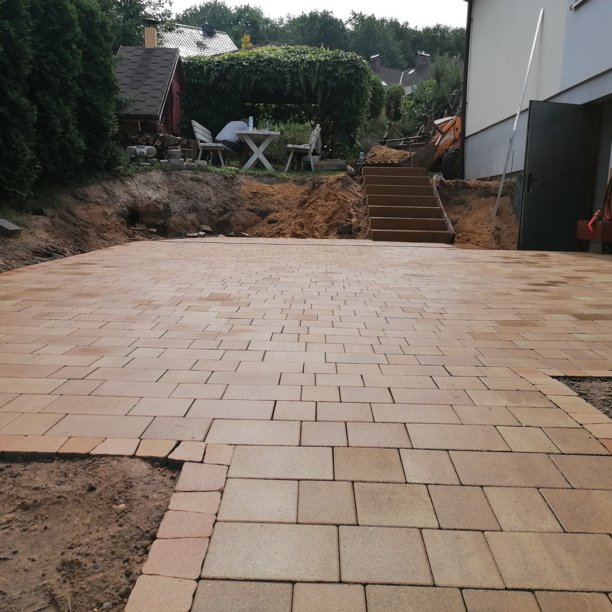 Układanie beżowej kostki brukowej na podjeździe, widoczne schody i ziemia do wyrównania, częściowo ułożona obwódka. Prace brukarskie w toku.