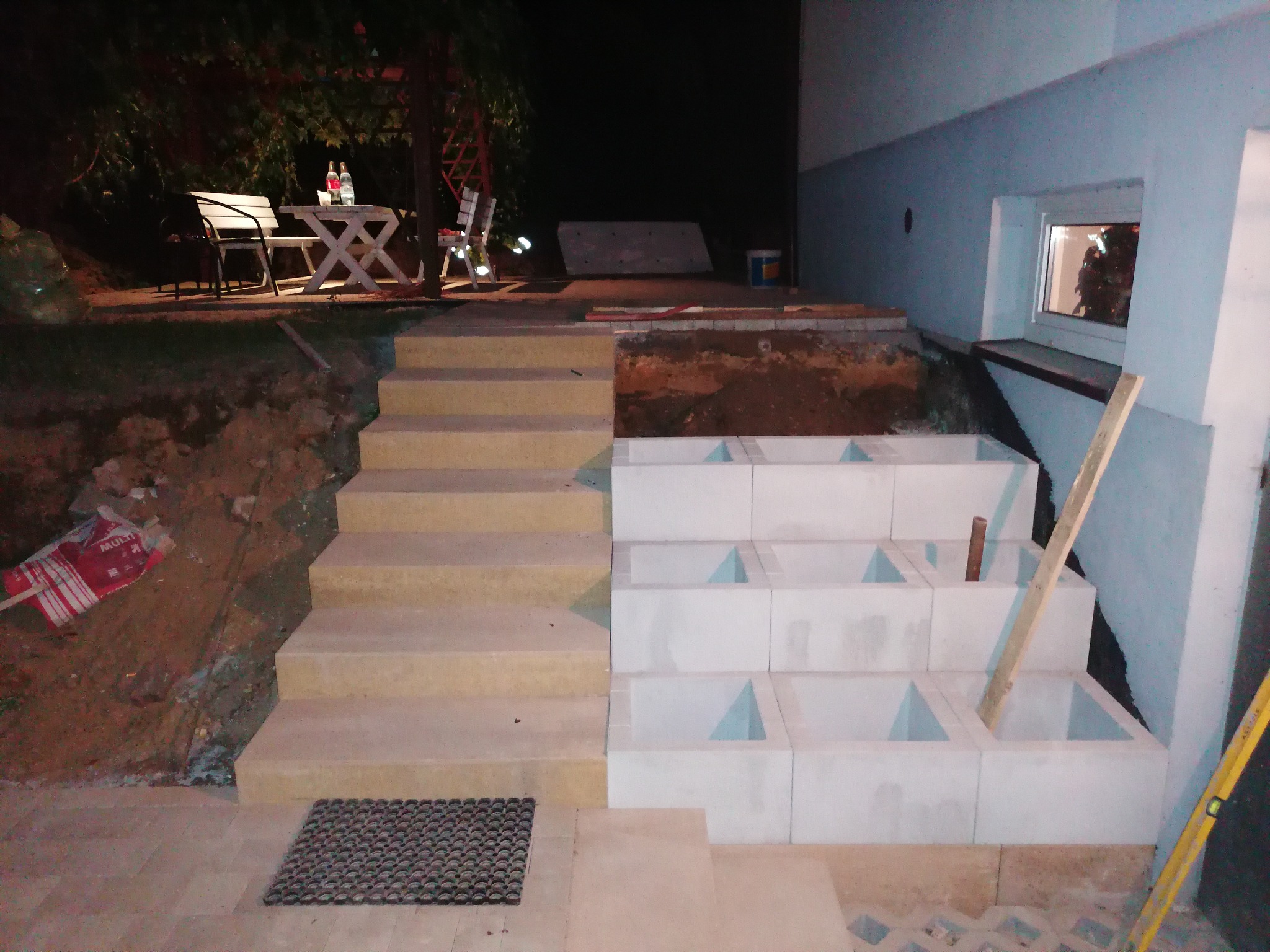 Budowa schodów zewnętrznych z bloczków betonowych i kostki, widoczne narzędzia i materiały budowlane, praca w toku, oświetlenie nocne.