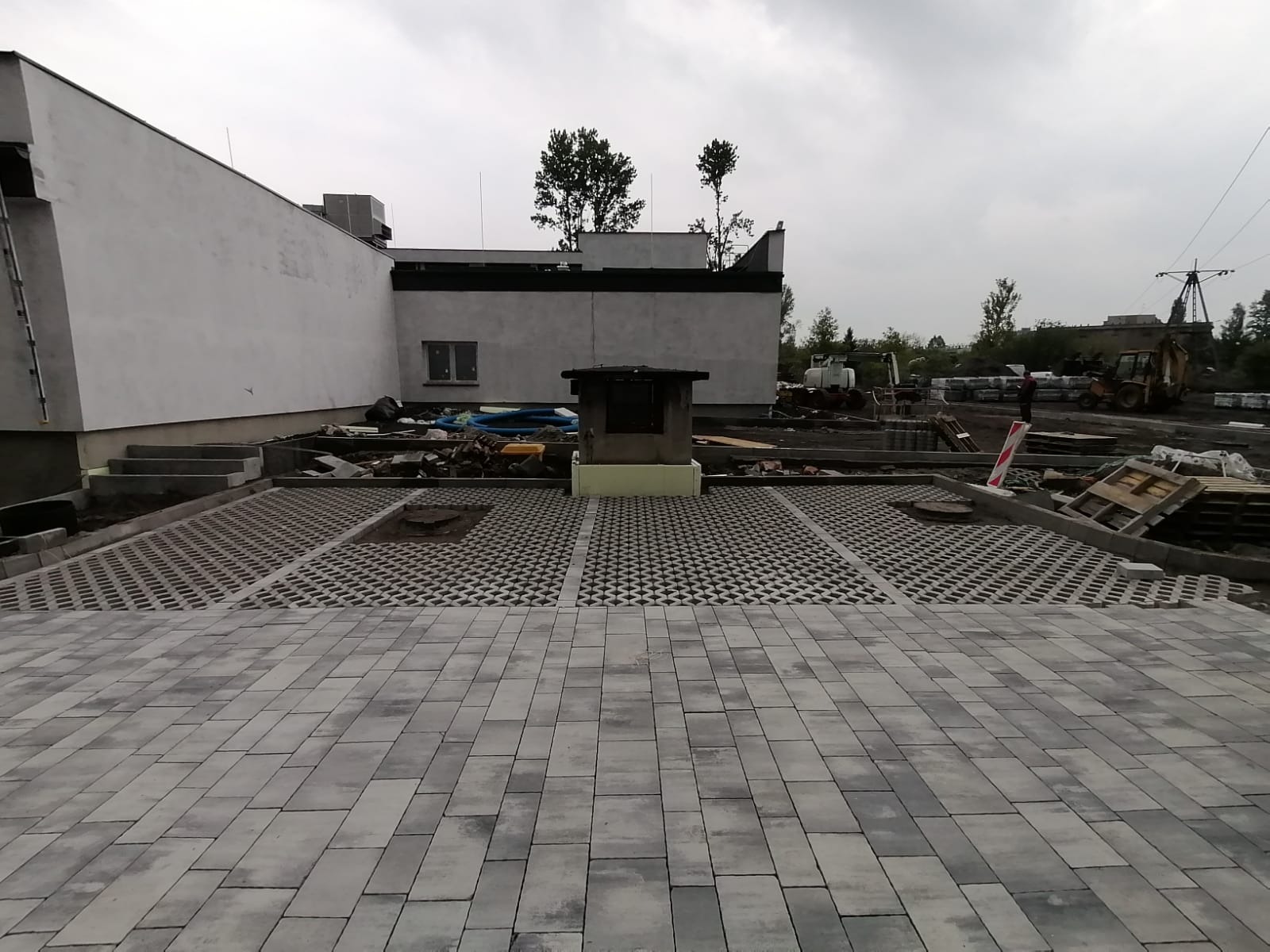 Fragment układania szarej kostki brukowej przed budynkiem, widoczne różne wzory ułożenia, palety z kostką w tle i maszyny budowlane. Prace w toku.