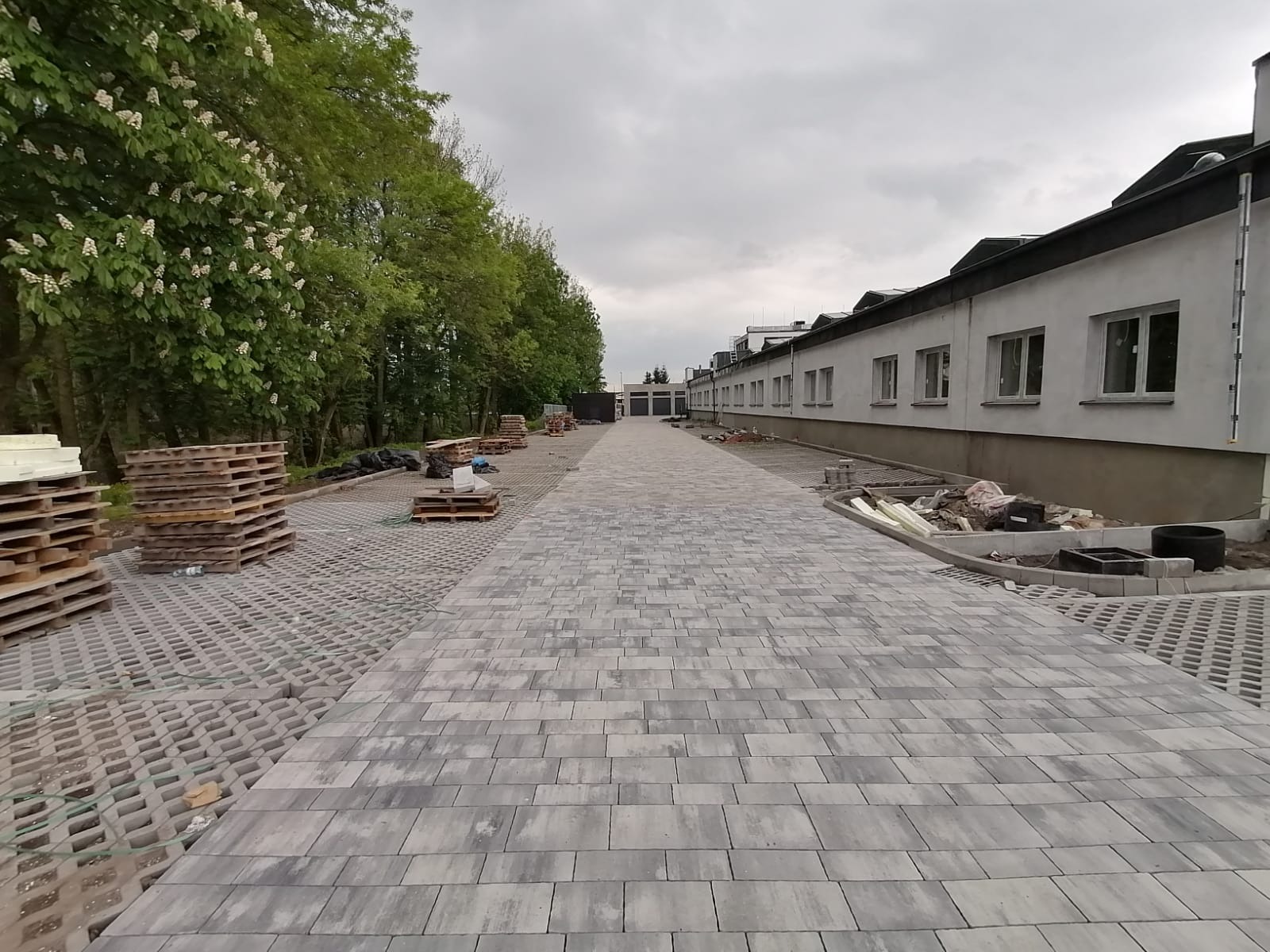 Szeroki, szary chodnik z kostki brukowej prowadzący wzdłuż budynku. Po lewej stronie palety z materiałami i drzewa. Widok w perspektywie, pochmurne niebo.