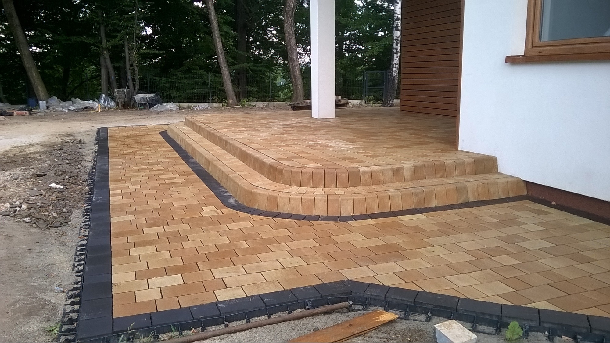 Ułożona kostka brukowa w odcieniach beżu i brązu z czarnym obramowaniem tworzy ścieżkę i schody przed budynkiem. Widoczne drzewa w tle.