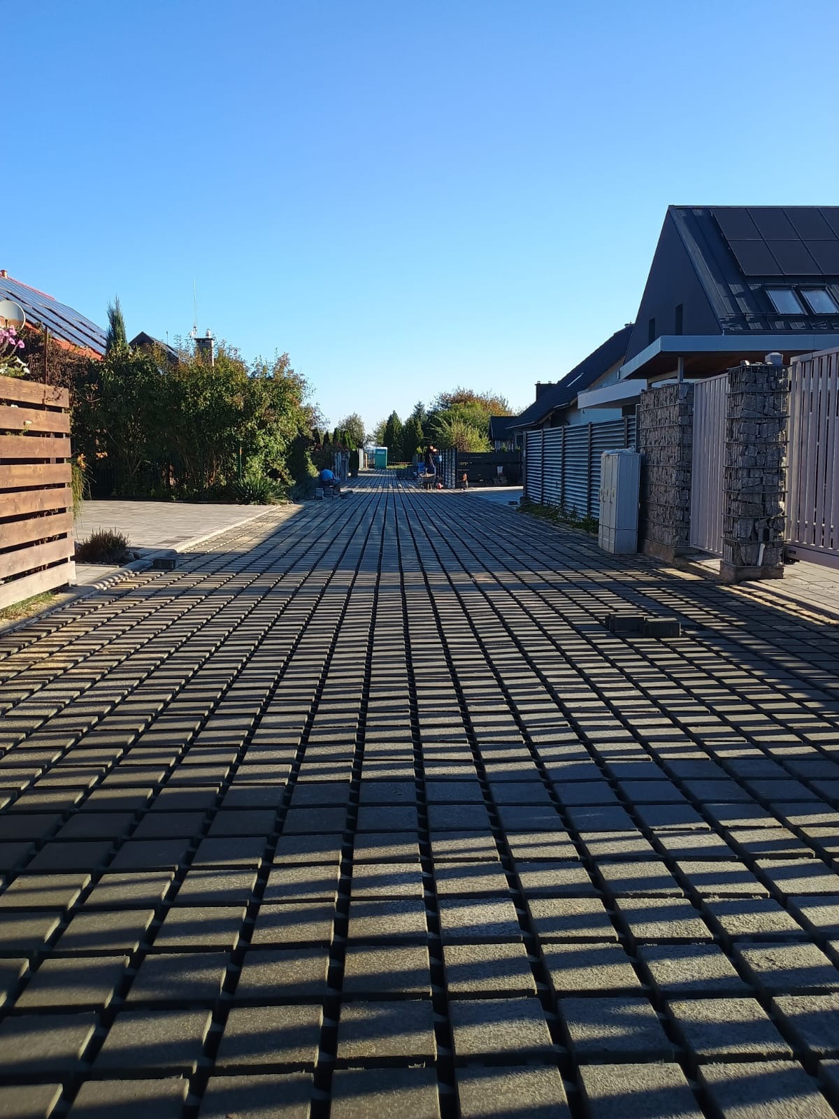 Długa, wybrukowana kostką droga dojazdowa do domów jednorodzinnych w słoneczny dzień. Widoczni pracownicy w oddali. Geometryczny wzór cieni na kostce.