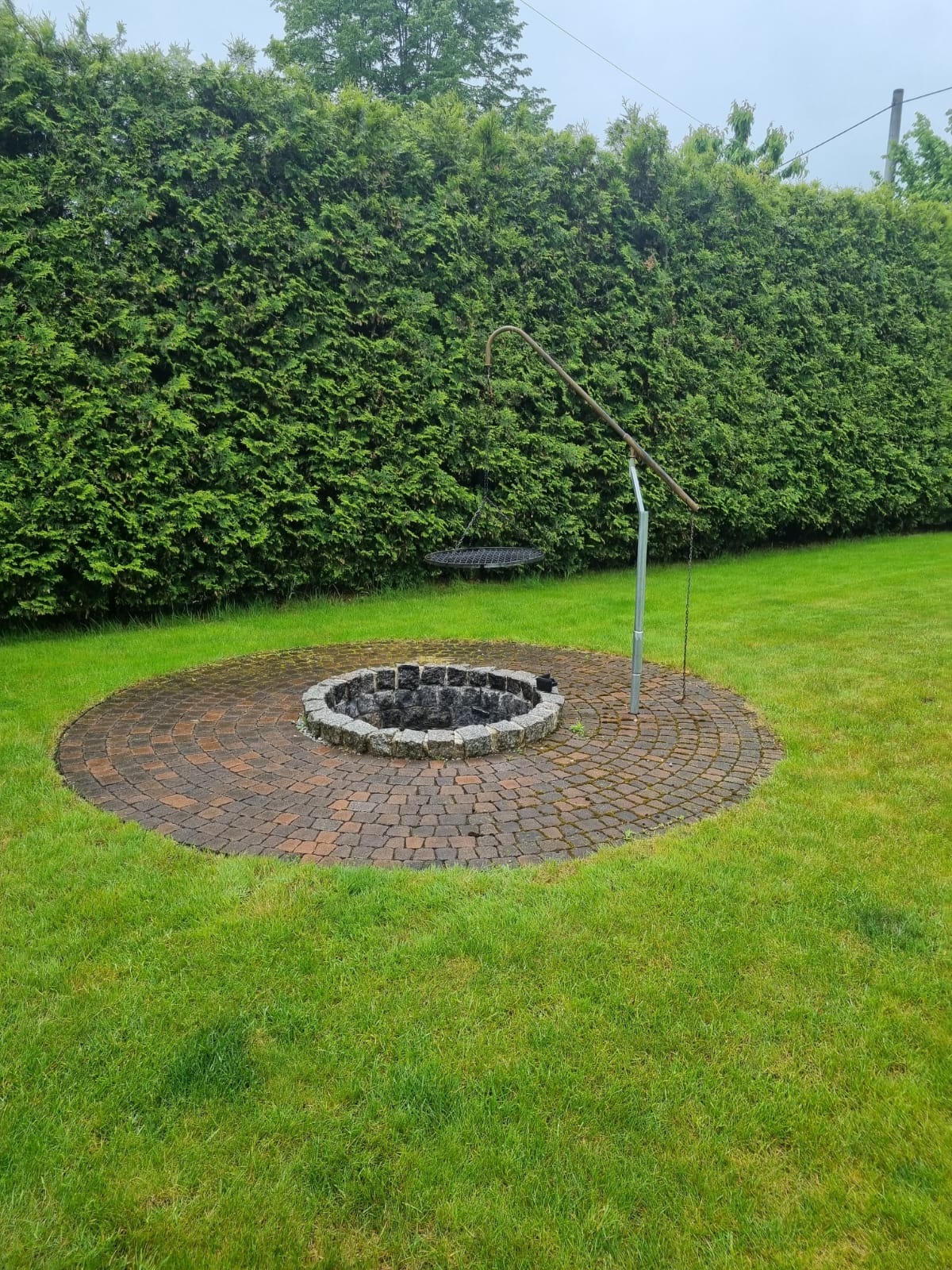 Ognisko z obudową z kostki granitowej, otoczone trawnikiem i żywopłotem. Na paleniskiem zawieszony ruszt. Widok z poziomu trawy, pochmurny dzień.