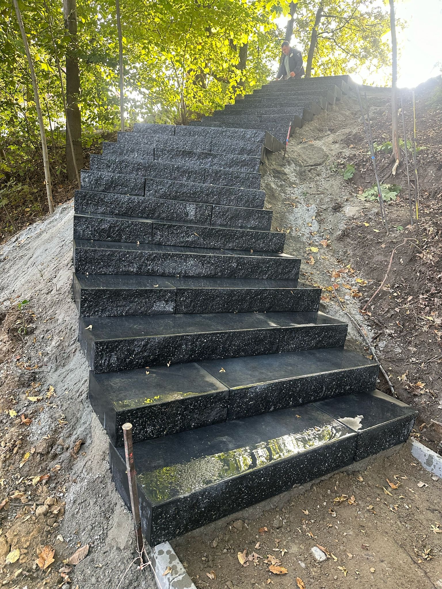 Granitowe schody na zboczu, prowadzące przez las. Widoczne czarne, błyszczące stopnie i surowe, ziemne otoczenie. U góry sylwetka osoby.