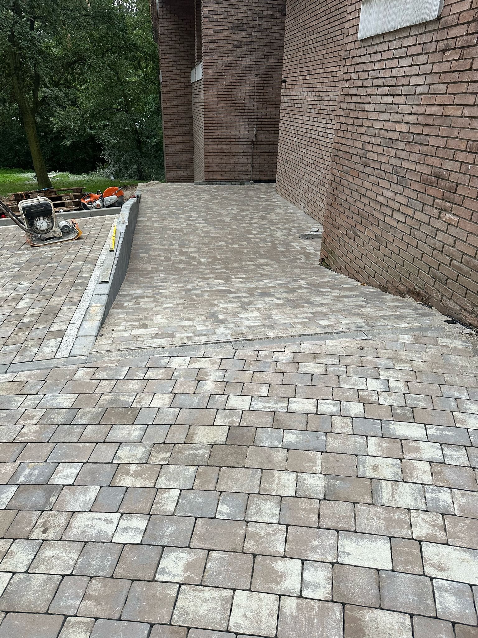 Ułożona kostka brukowa w odcieniach szarości i beżu, tworząca ścieżkę przy ceglanym budynku. Widoczny sprzęt budowlany i fragment zieleni w tle.