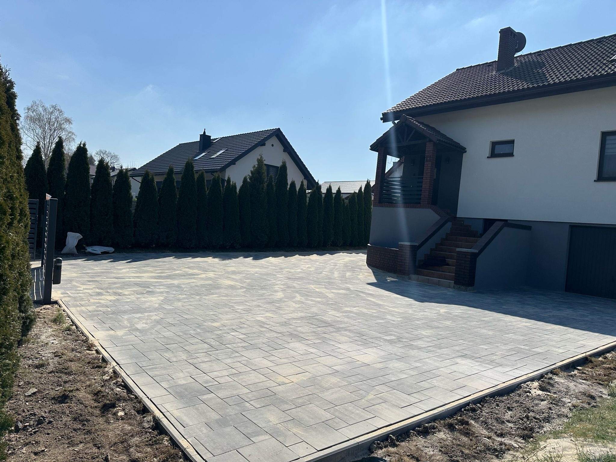 Szeroki podjazd z szarej kostki brukowej ułożonej we wzór prostokątów, obramowany obrzeżem. W tle dom z brązowym dachem i rząd tuj. Słoneczny dzień.