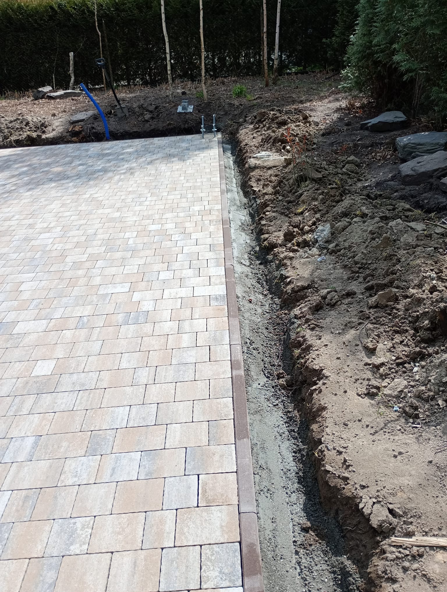 Fragment układania kostki brukowej beżowo-szarej. Widoczna krawędź z ciemniejszych kostek i wykop z ziemią. W tle zieleń, narzędzia i elementy instalacji.