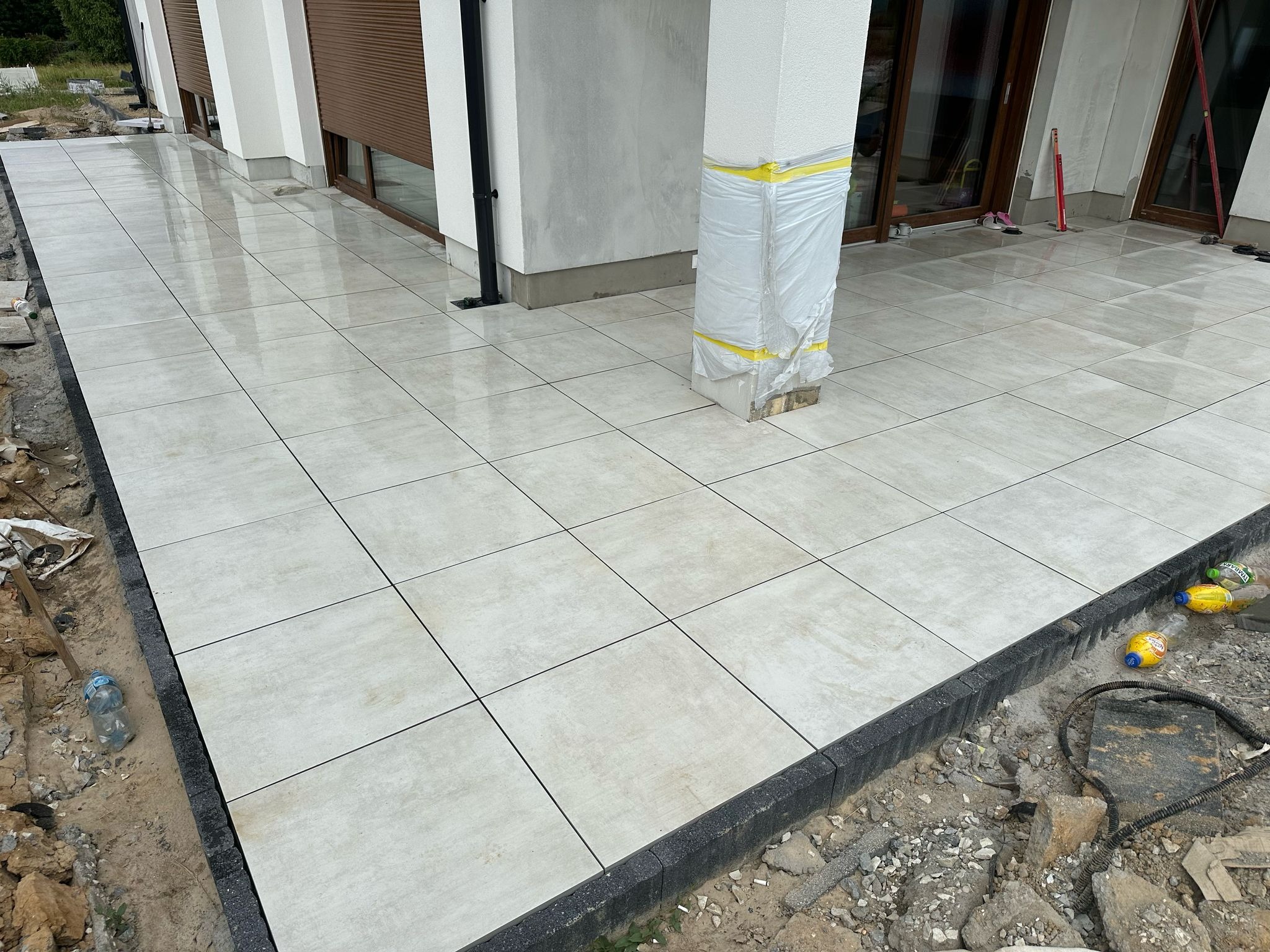Ułożona jasna, kwadratowa terakota na zewnątrz budynku, obramowana ciemną kostką. Część powierzchni jeszcze nieukończona, widoczne narzędzia i butelki.