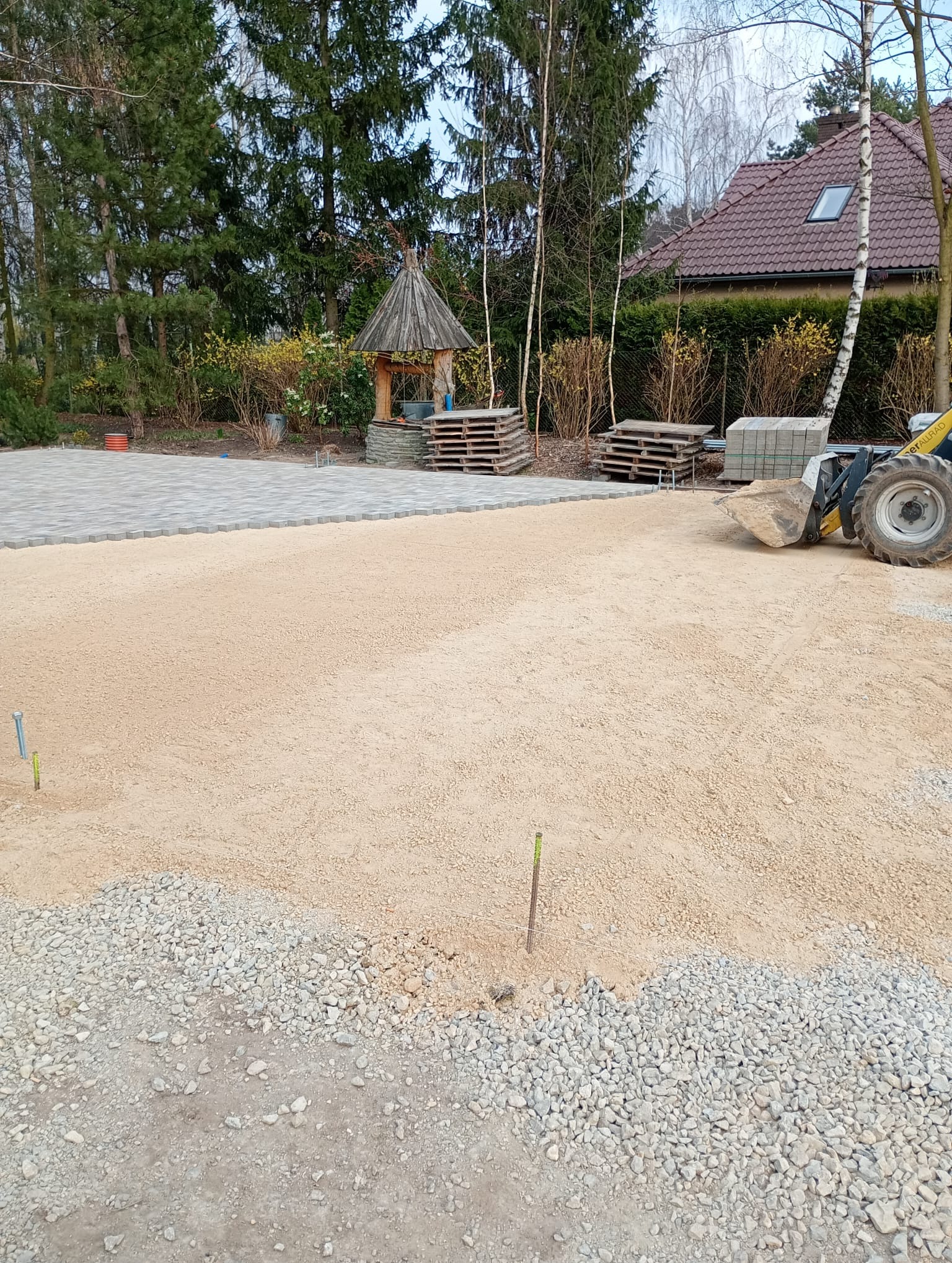Etap przygotowania terenu pod kostkę brukową: wyrównany piasek, częściowo ułożona kostka, palety z kostką i mała koparka w tle, drzewa i altana.