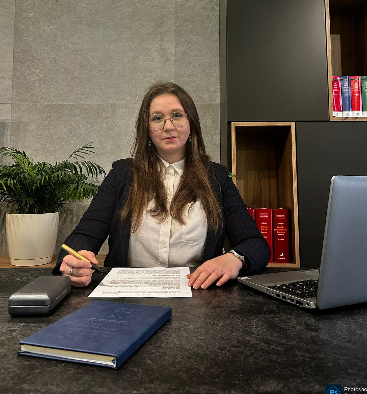 Kobieta w biurze, podpisuje dokument złotym piórem, laptop i książki w tle. Elegancki wystrój, profesjonalne otoczenie.