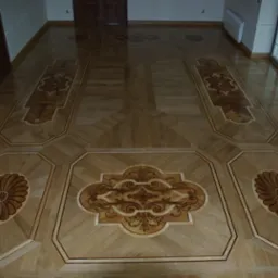 Wzorzysta podłoga drewniana z intarsjami o motywach kwiatowych i ornamentacyjnych, ułożona w przestronnym wnętrzu z widocznymi drzwiami i oknami.