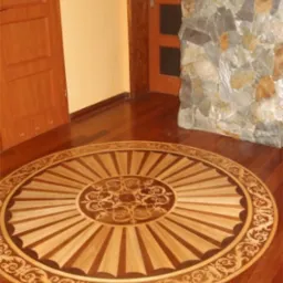 Drewniana podłoga z dekoracyjnym, okrągłym wzorem parkietu w stylu klasycznym, otoczona panelami w kolorze ciemnego drewna, widoczne drzwi i fragment ściany z kamienia.