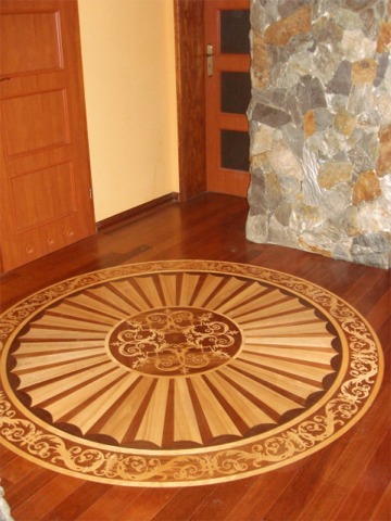 Drewniana podłoga z dekoracyjnym, okrągłym wzorem parkietu w stylu klasycznym, otoczona panelami w kolorze ciemnego drewna, widoczne drzwi i fragment ściany z kamienia.