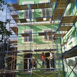 BUILD WEST-Yaroslav Sysoiev - Zielona elewacja domu w trakcie malowania, widoczna z rusztowania, z pracownikami w trakcie pracy. Puszki z farbą na rusztowaniu, słoneczny dzień.