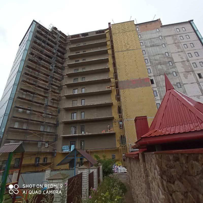 Wielopiętrowy budynek w trakcie prac elewacyjnych: ocieplanie fragmentów, rusztowania, widoczne różne etapy prac. Ujęcie od dołu, fragment czerwonego dachu w kadrze.