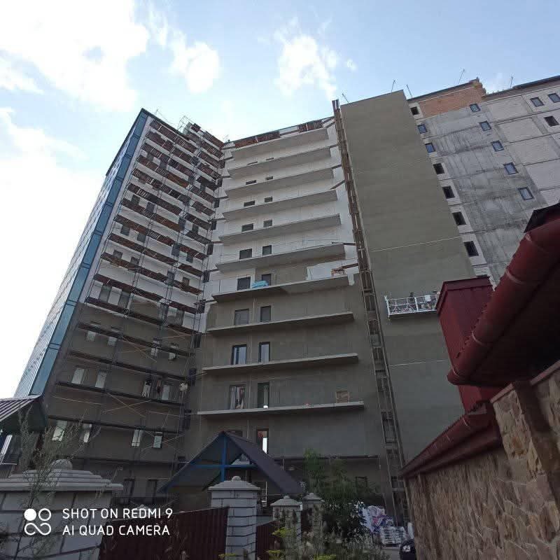 Wielopiętrowy budynek w trakcie prac elewacyjnych. Widoczne rusztowania, fragment ocieplonej ściany i niezabezpieczone balkony. Ujęcie z dołu, pochmurne niebo.