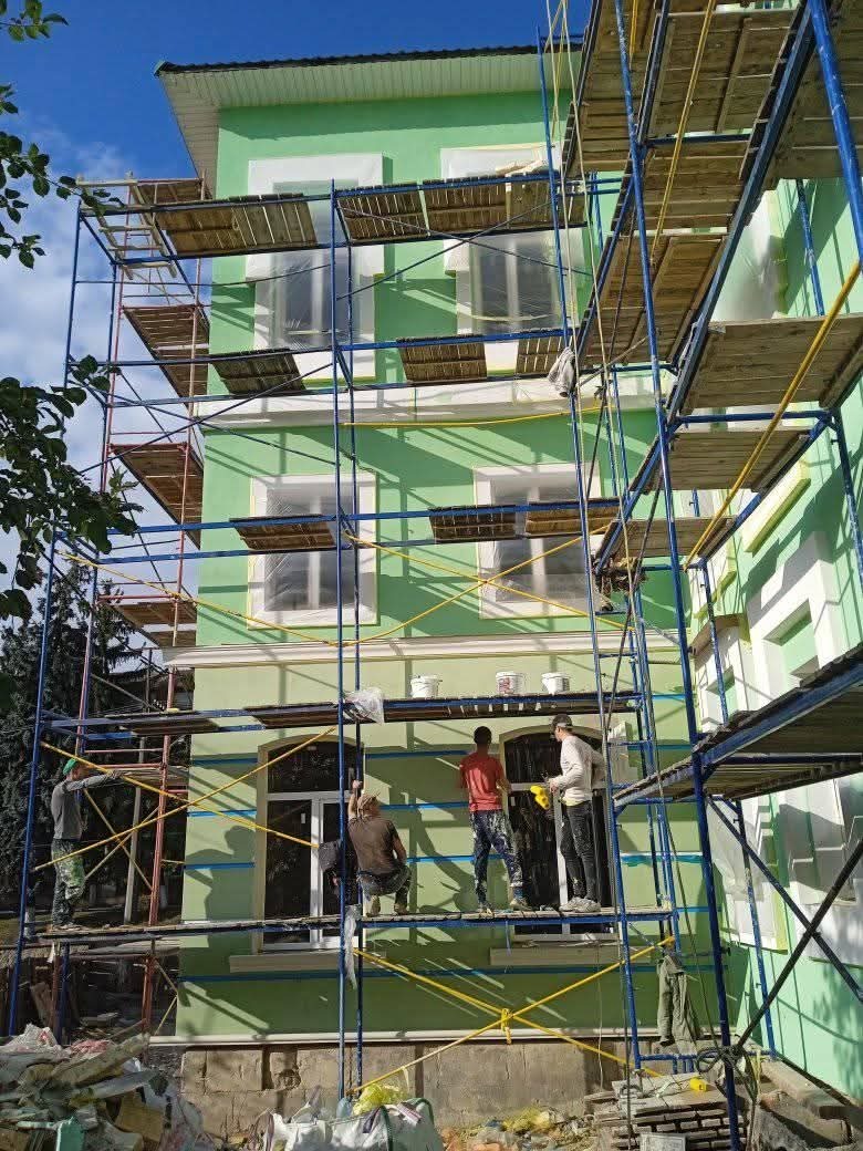 Zielona elewacja domu w trakcie malowania, widoczna z rusztowania, z pracownikami w trakcie pracy. Puszki z farbą na rusztowaniu, słoneczny dzień.