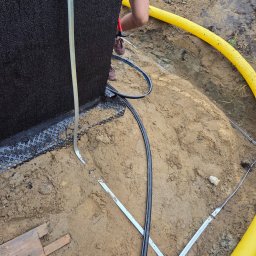 AN InstalTech Arkadiusz Nagórski - Ocieplenie fundamentu budynku z użyciem czarnej membrany i żółtej rury drenażowej, widoczne elementy mocujące i wykop ziemny.