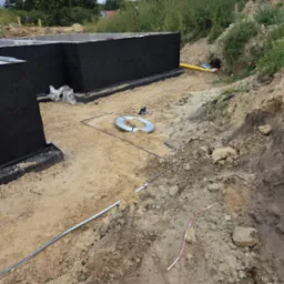 Fundamenty domu z izolacją przeciwwilgociową, widoczne rury i elementy instalacji w wykopie. Prace przygotowawcze do dalszych etapów budowy.