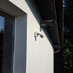 AN InstalTech Arkadiusz Nagórski - Zewnętrzny reflektor LED zamontowany na elewacji domu z oknem i drewnianym okapem dachu. Widoczny fragment rynny i zieleń w tle.