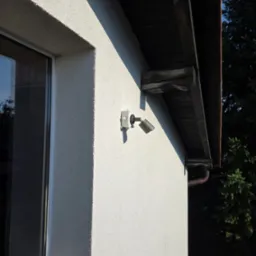 Zewnętrzny reflektor LED zamontowany na elewacji domu z oknem i drewnianym okapem dachu. Widoczny fragment rynny i zieleń w tle.