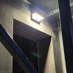 AN InstalTech Arkadiusz Nagórski - Zamontowana lampa zewnętrzna na szorstkiej ścianie obok otworu drzwiowego, z widoczną kratką sufitową i elementami konstrukcyjnymi, oświetlona od góry.