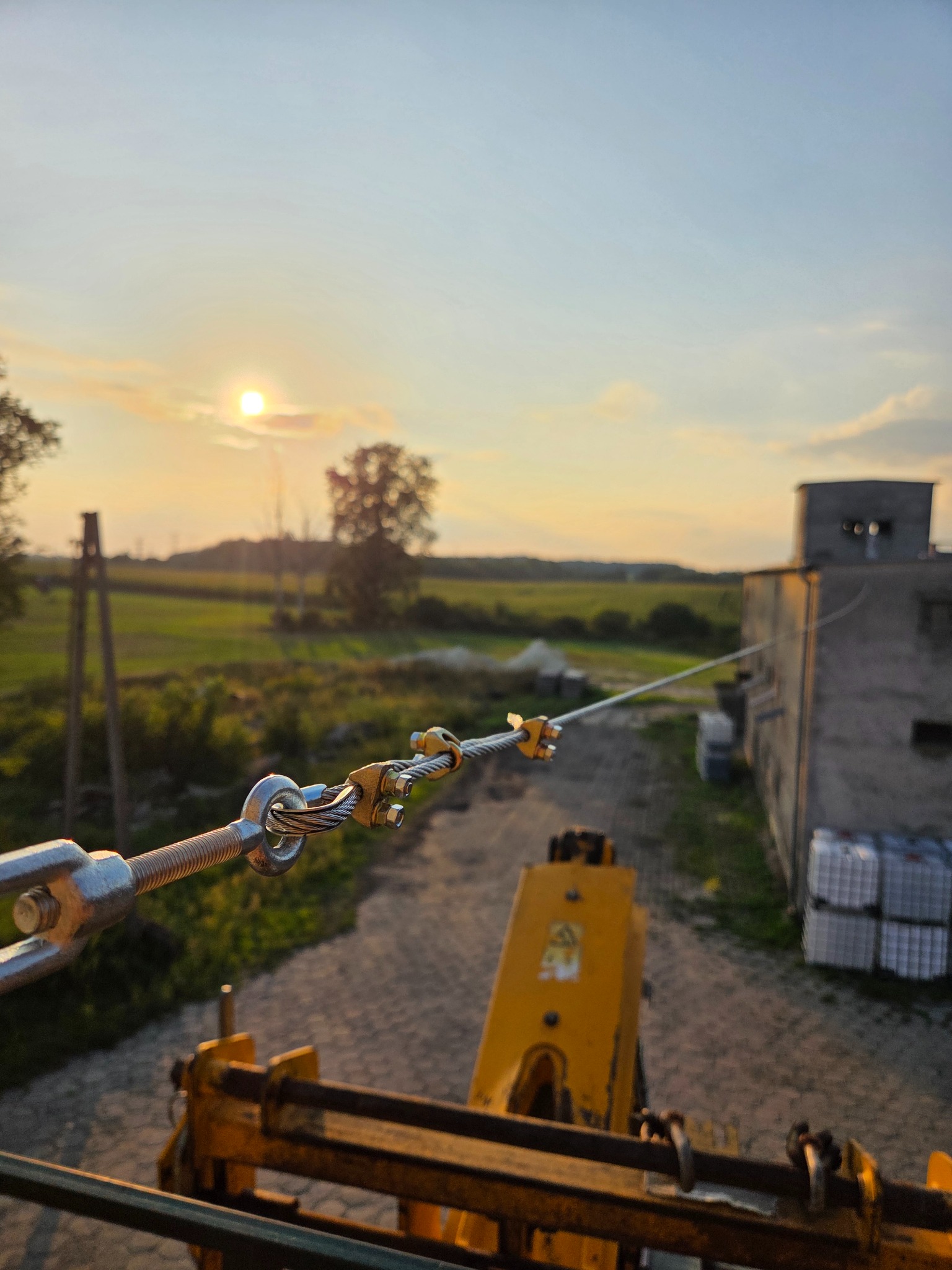 Napinacz liny stalowej z widokiem na zachodzące słońce i polny krajobraz. Elementy metalowe na pierwszym planie, budynek w tle po prawej stronie.