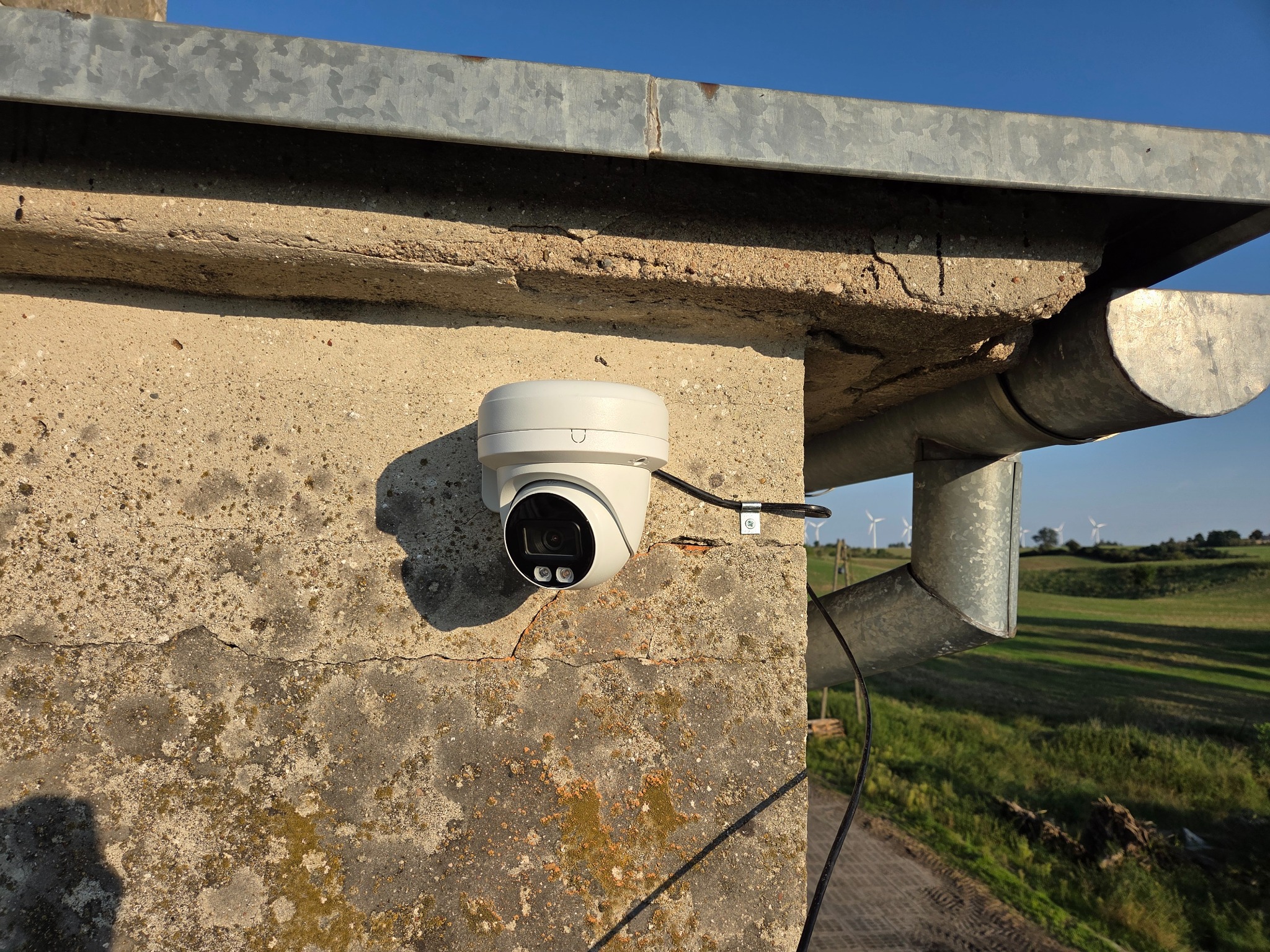 Biała kamera monitoringu kopułkowa przymocowana do betonowej ściany budynku z widoczną rynną i krajobrazem w tle, instalacja zewnętrzna, zbliżenie.