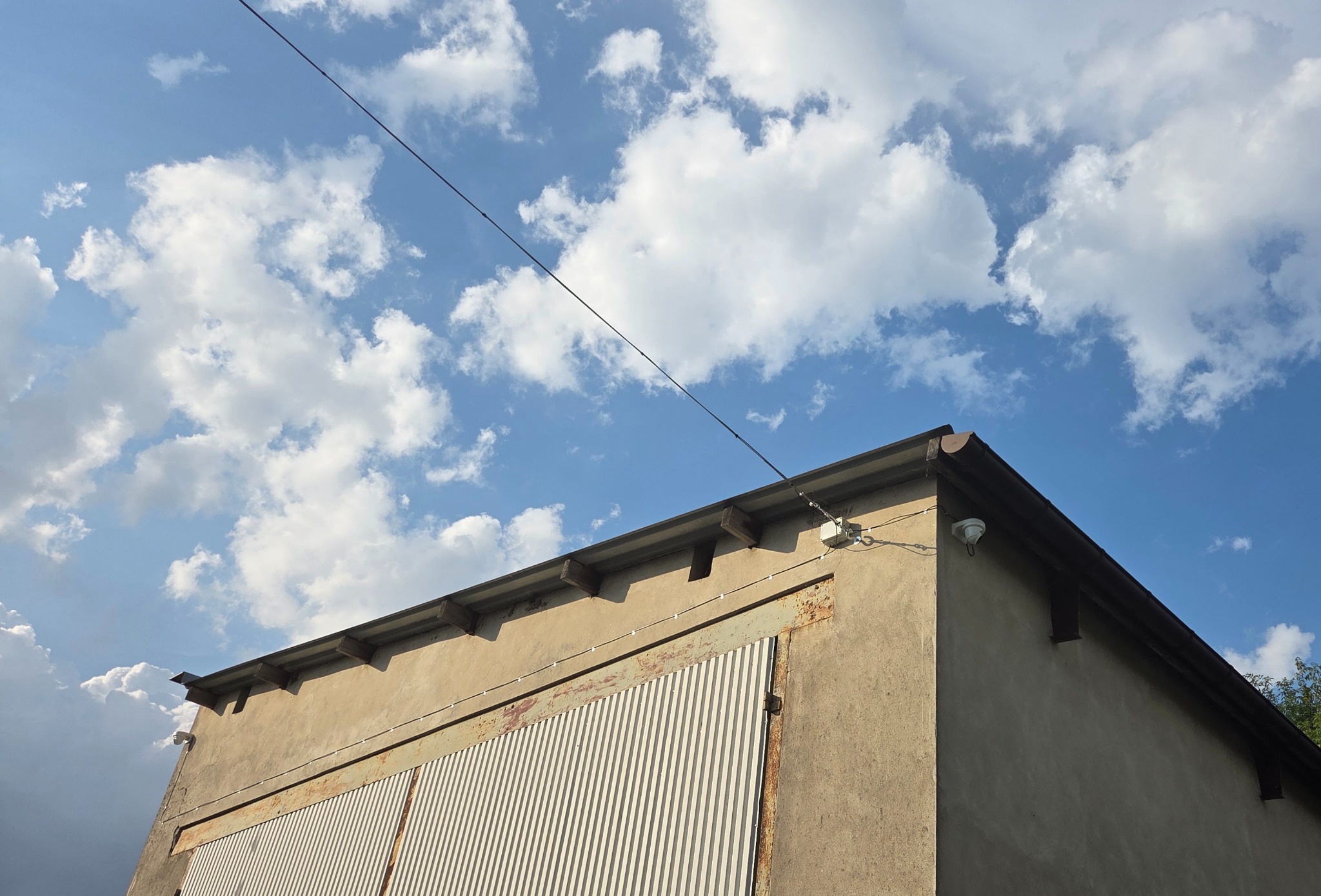 Kamera monitoringu na rogu budynku z metalową bramą, na tle błękitnego nieba z chmurami. Widoczne przewody elektryczne.