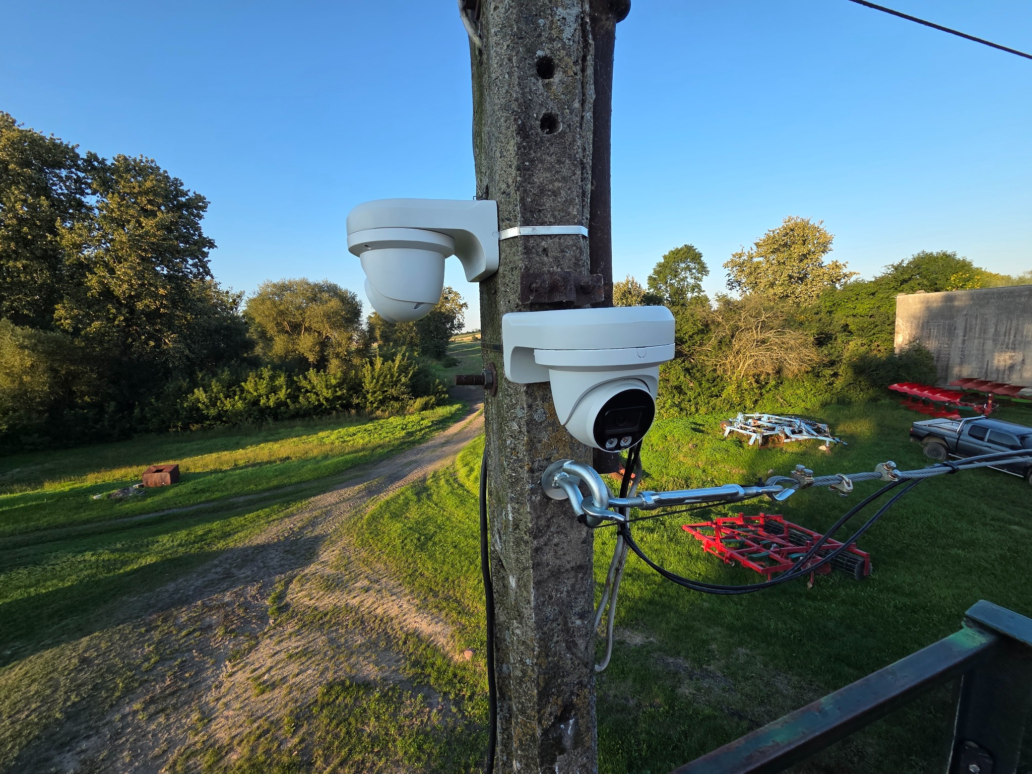 Dwie białe kamery monitoringu kopułkowe zamontowane na betonowym słupie, widok z góry na wiejski krajobraz z polami i drzewami w tle.