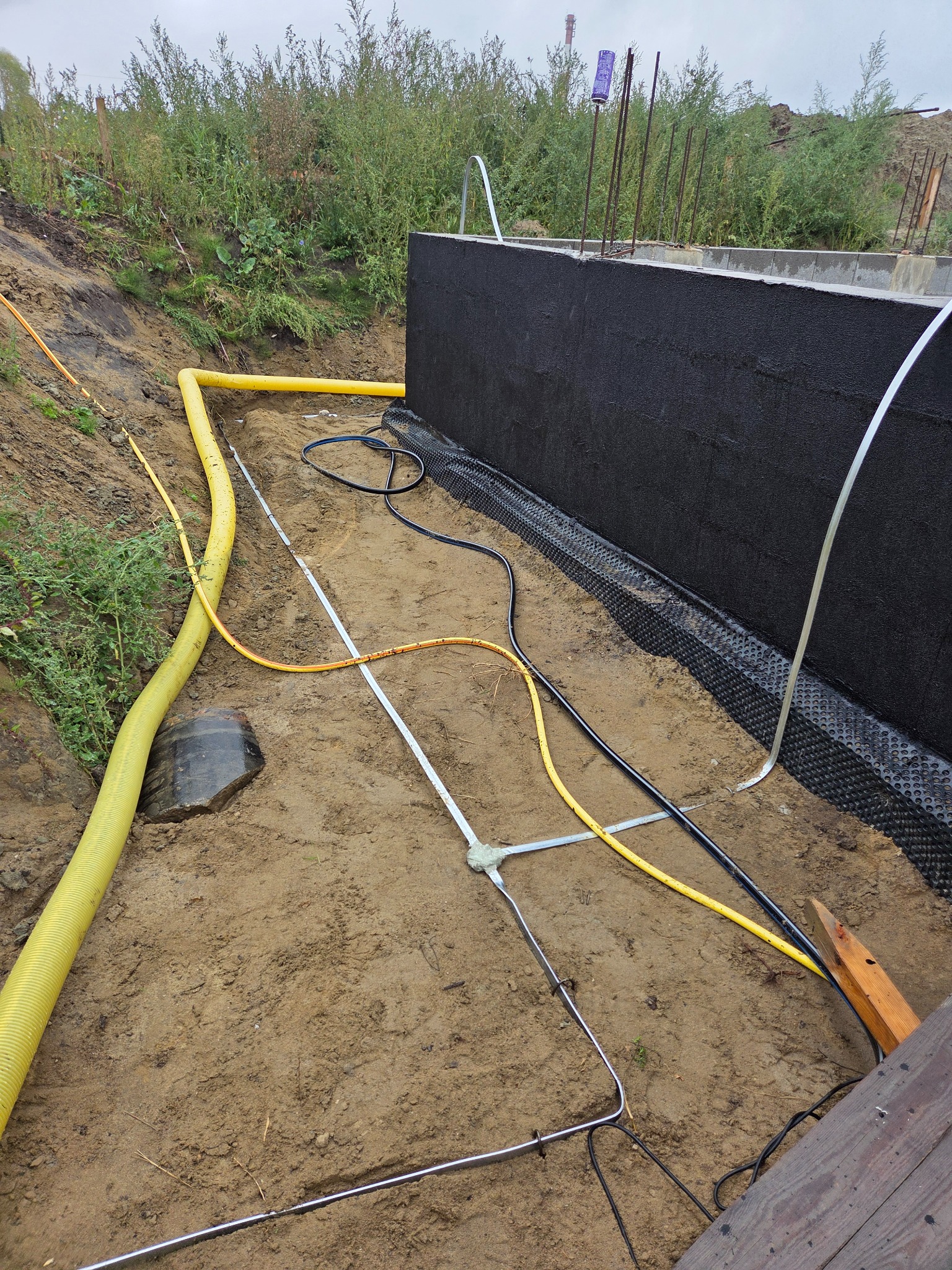Izolacja fundamentów z widocznymi rurami instalacyjnymi w wykopie. Prace ziemne, zabezpieczenie hydroizolacyjne i instalacja rur w gruncie.