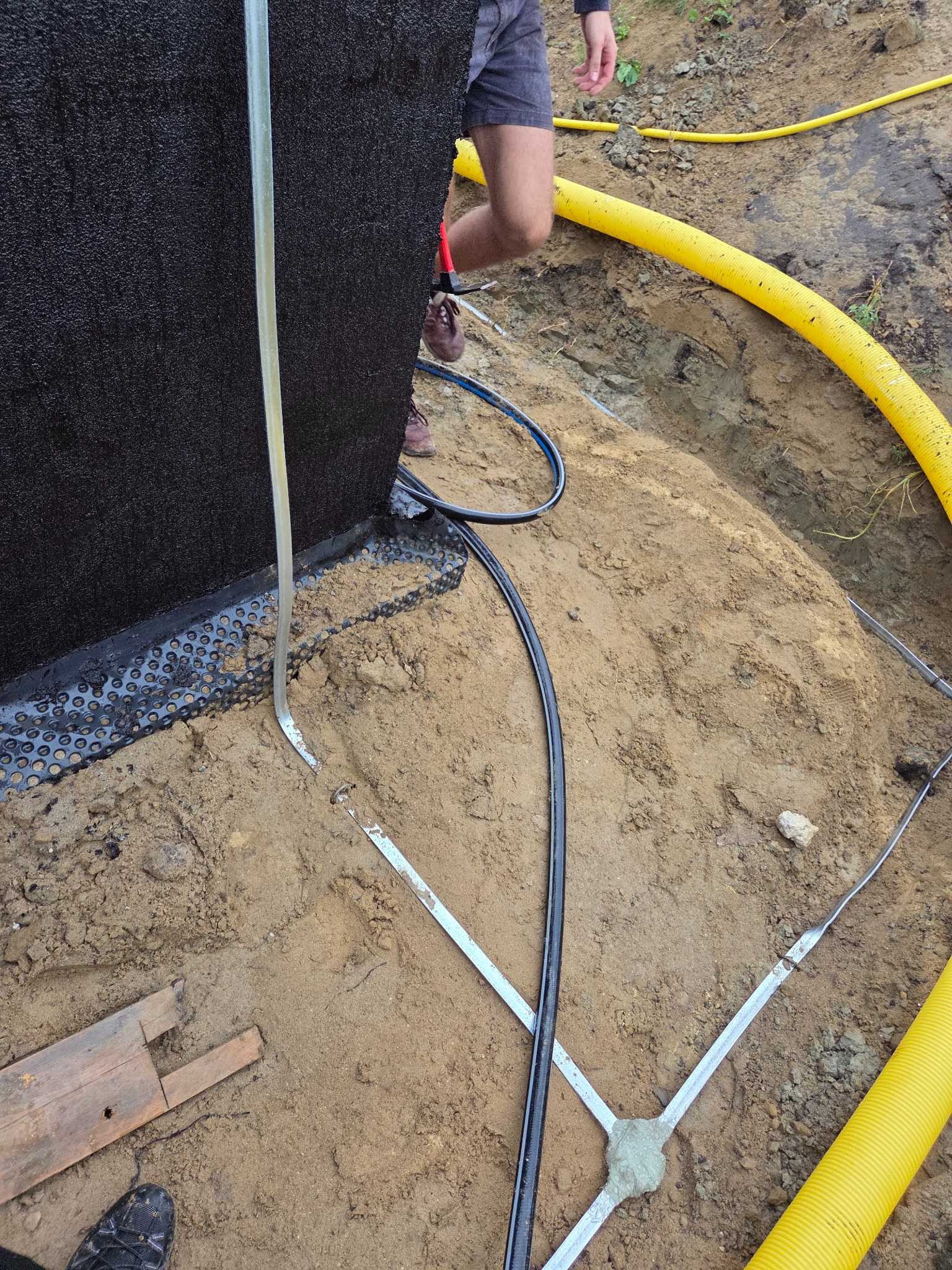 Ocieplenie fundamentu budynku z użyciem czarnej membrany i żółtej rury drenażowej, widoczne elementy mocujące i wykop ziemny.